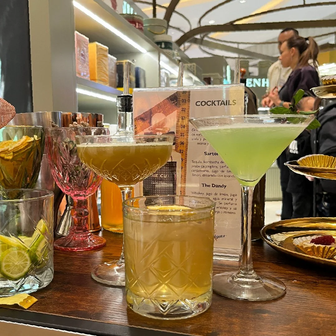 variedad de cócteles en una barra, incluyendo uno verde y uno amarillo, con algunos ingredientes y utensilios de bar en el fondo, y personas en un ambiente de bar o restaurante.