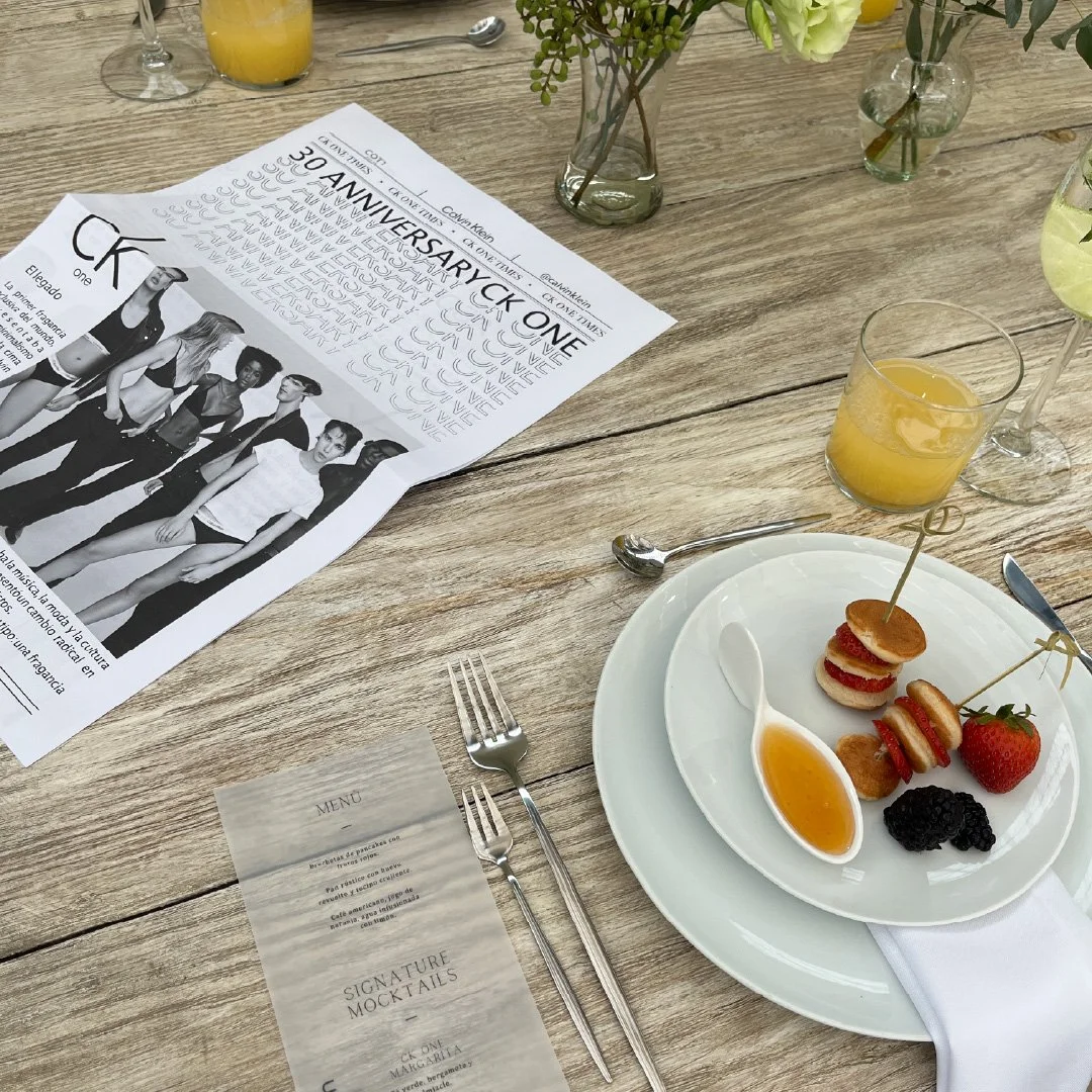 Mesa de madera con un plato de aperitivos y frutas, un vaso con jugo de naranja, y un folleto de celebración del 30 aniversario. También hay un menú, cubiertos y varios vasos con bebidas.