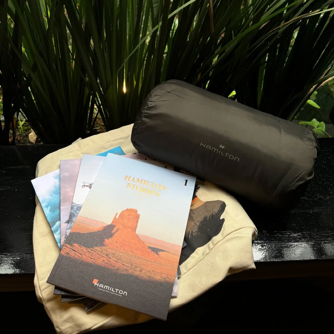 Revista de historias de Hamilton, una bolsa de tela, un cilindro negro con la marca Hamilton, y un par de libros con un fondo de plantas verdes.