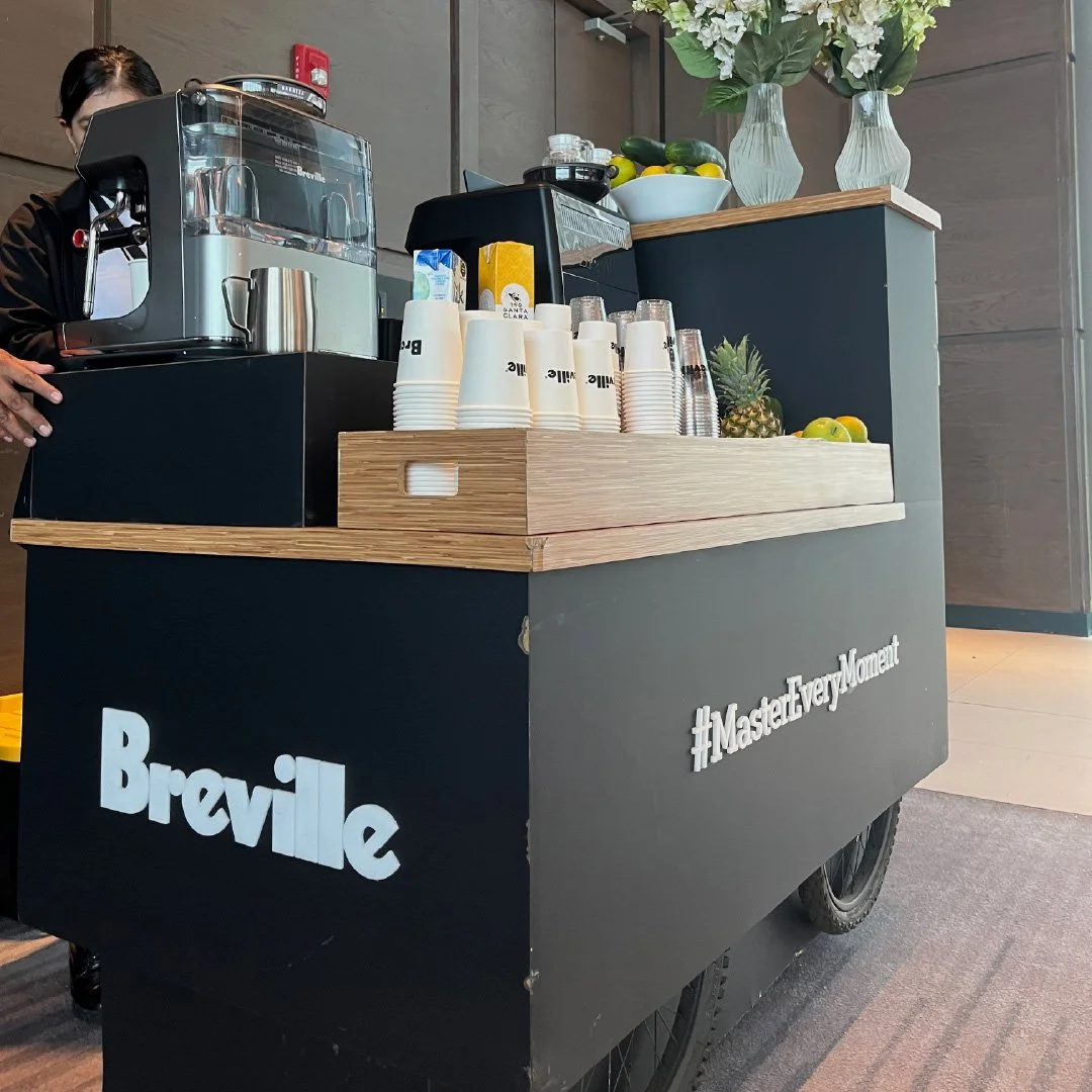 Carrito de bebidas de café con máquina de espresso, vasos desechables, piña, y frutas variadas, decorado con flores en jarrones