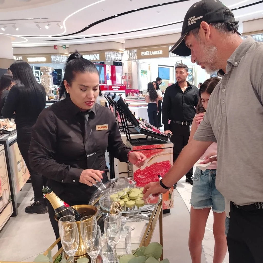 Una mujer con uniforme de tienda atiende a un cliente en un puesto con macarons verdes, botellas de vino y copas de vino en un centro comercial.