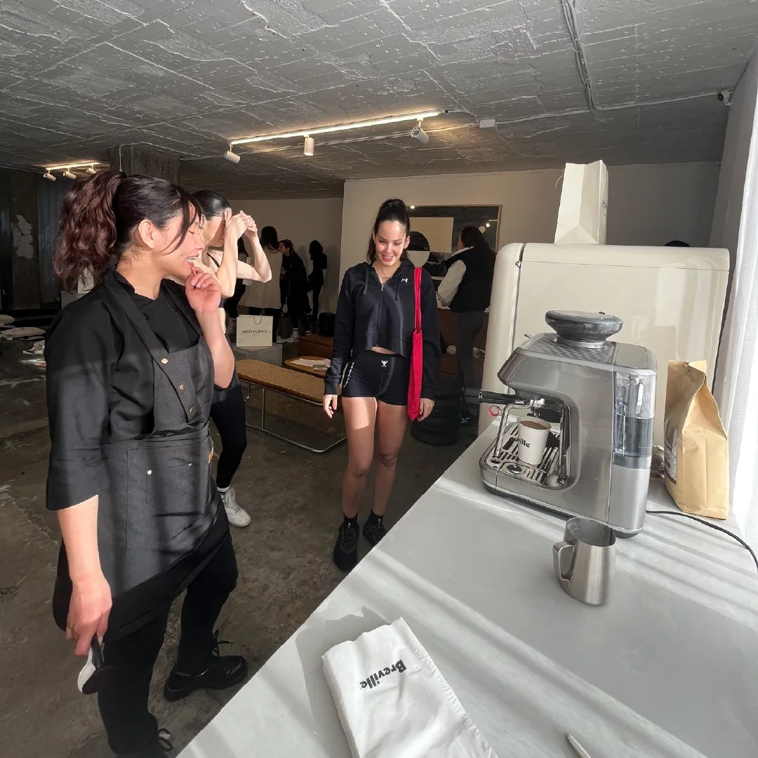 Un grupo de mujeres en una cafetería. Una mujer con uniforme de camarera frente a una máquina de café. Otra mujer con ropa deportiva y una mochila roja, sonriendo.