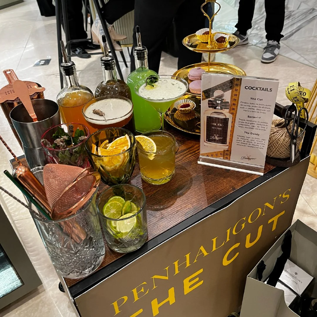 Mostrador con varias bebidas en vasos y botellas, una bandeja con macarons, un cartel con la lista de cócteles y algunos utensilios de bar.