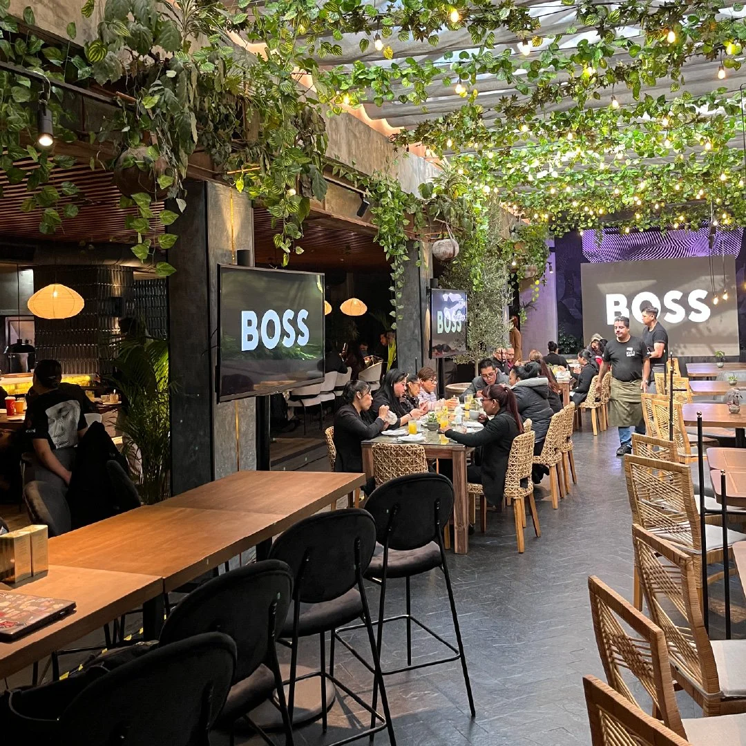 Restaurante con clientes sentados en mesas de madera y sillas de mimbre. Pantallas grandes con la palabra 'BOSS' en la pared del fondo, decoración con plantas y luces colgantes.