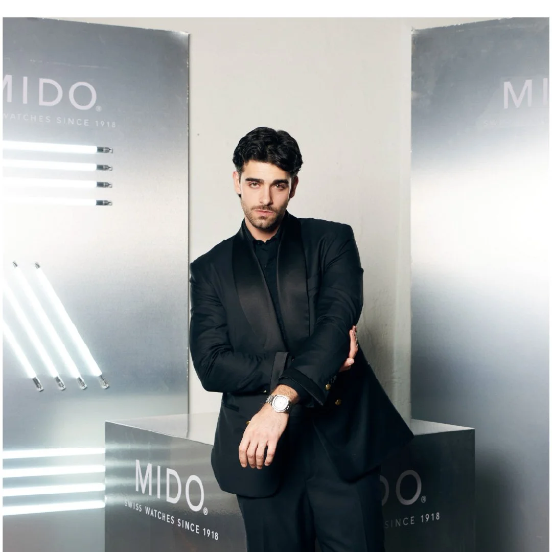 Hombre joven con barba, cabello oscuro y ropa formal negra, posa con los brazos cruzados en un entorno moderno y elegante, con anuncios de relojes MIDO en el fondo.