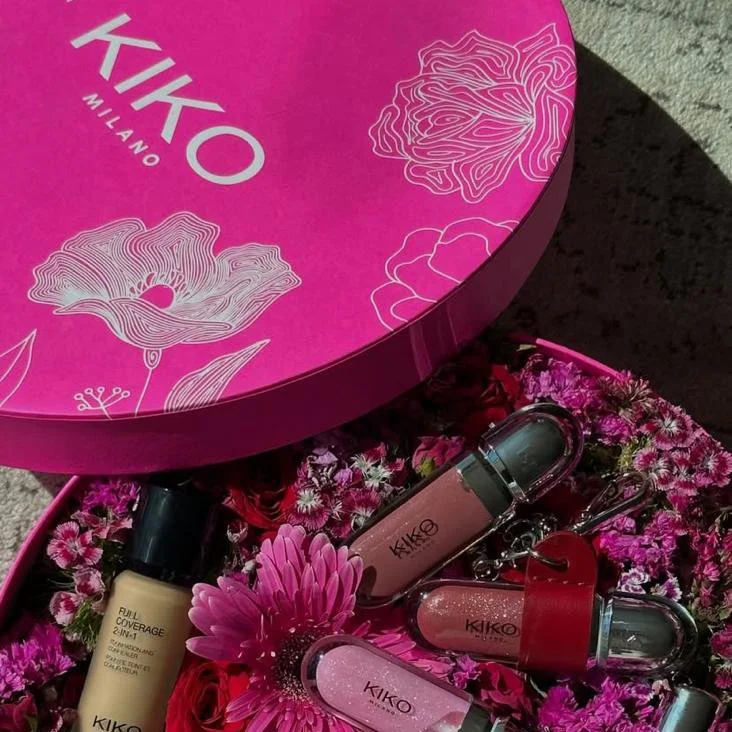 Caja de regalo circular de color rosa con el logo 'KIKO MILANO' y dibujos de flores blancas. Dentro hay varios productos de maquillaje y cosméticos rodeados de flores moradas y rosas.