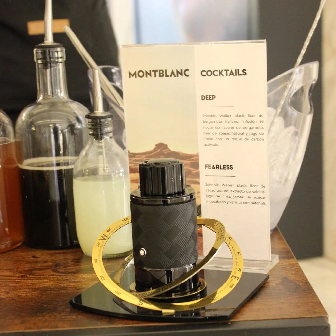 Varios frascos con líquidos, una caja con recetas de cócteles Montblanc y un vaso con hielo en una mesa de madera, con una brújula decorativa y una tarjeta con descripciones de bebidas. 