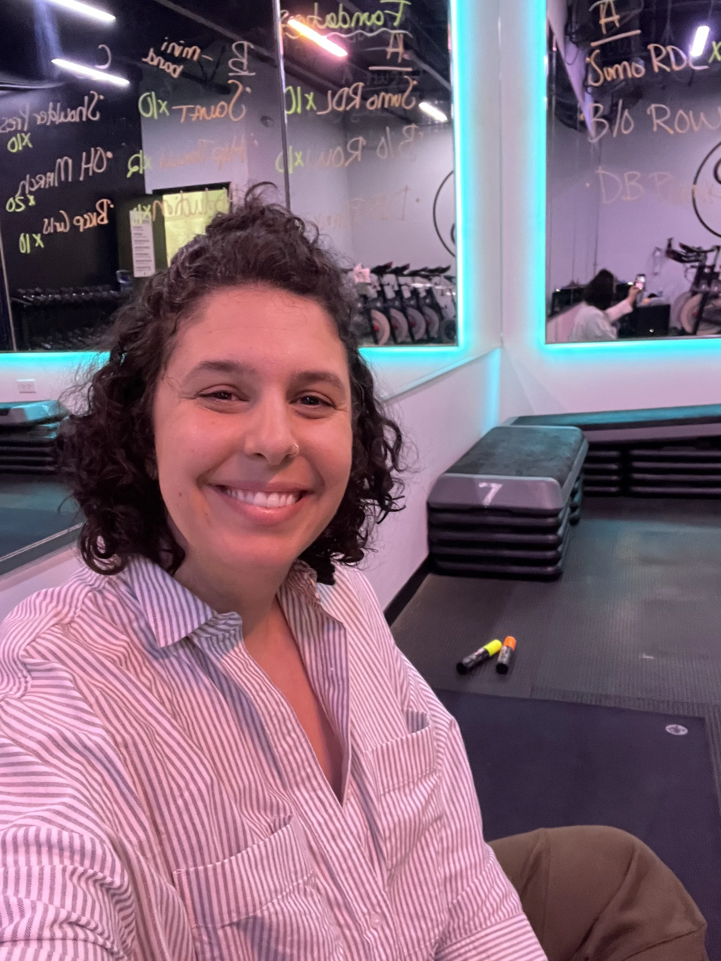 Dr. Grace is sitting in a gym, with mirrors on all walls. They are smiling into the camera, wearing a light gray stripped button up.