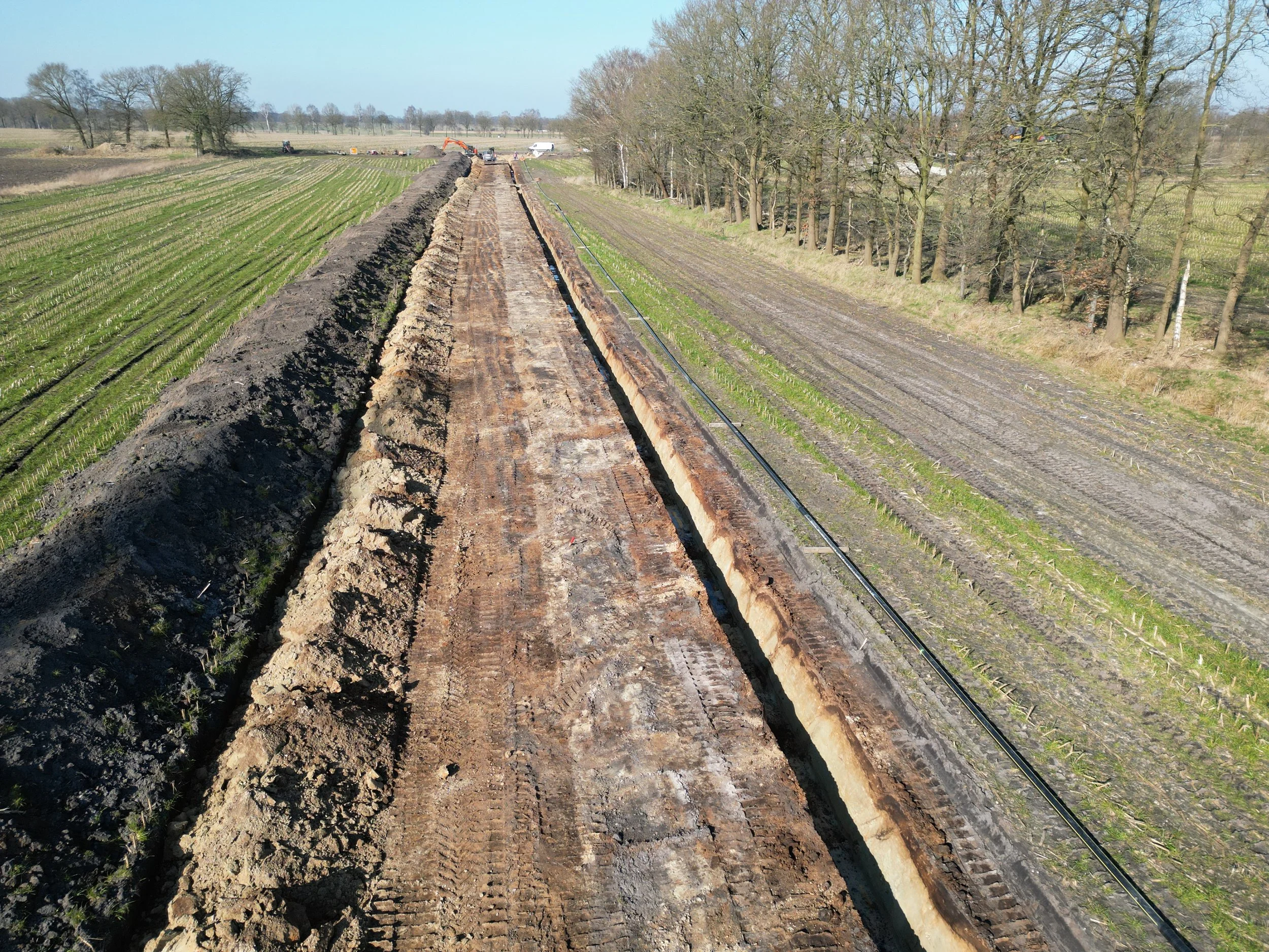 Bauarbeiten an einer langen, tiefen Baugrube in einer landwirtschaftlichen Umgebung mit Baufahrzeugen im Hintergrund.