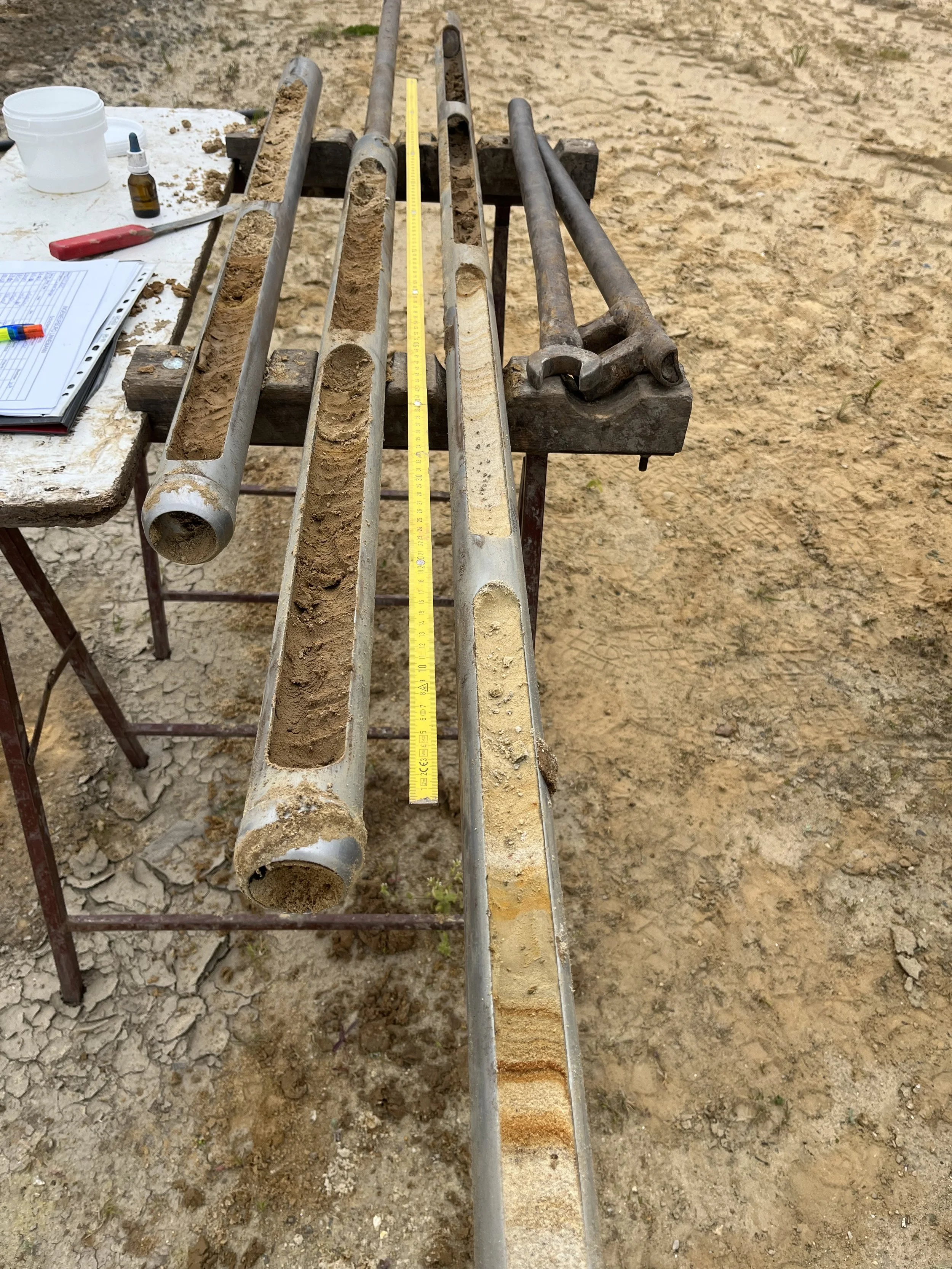 Lange Metallrohre und zwei große Schraubenschlüssel auf einem Werkbank im Freien, mit Messleiter und Werkzeugen auf der Seite.