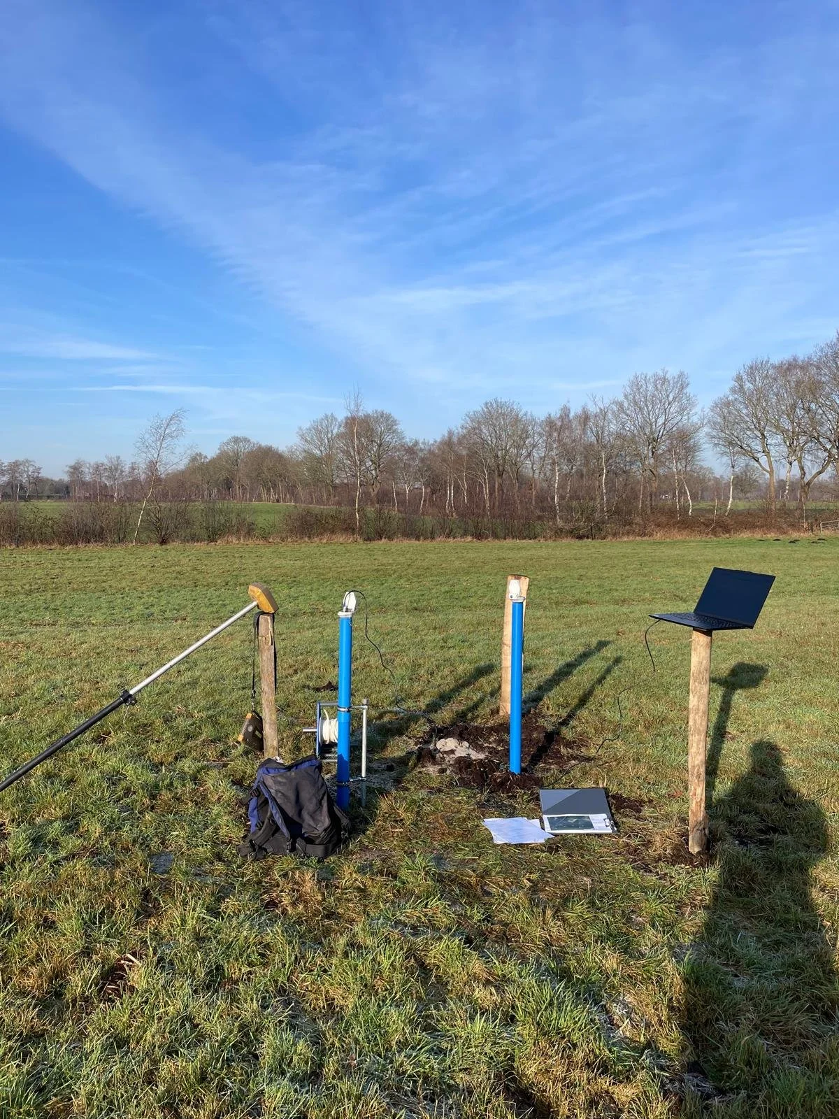 Ein offenes Feld mit drei blaue Rohrbrunnen, einem Laptop auf einem Holzpfosten, eine Tasche, und Wissenschaftliche Geräte, die auf den Boden verteilt sind, unter einem blauen Himmel mit vereinzelten Wolken.