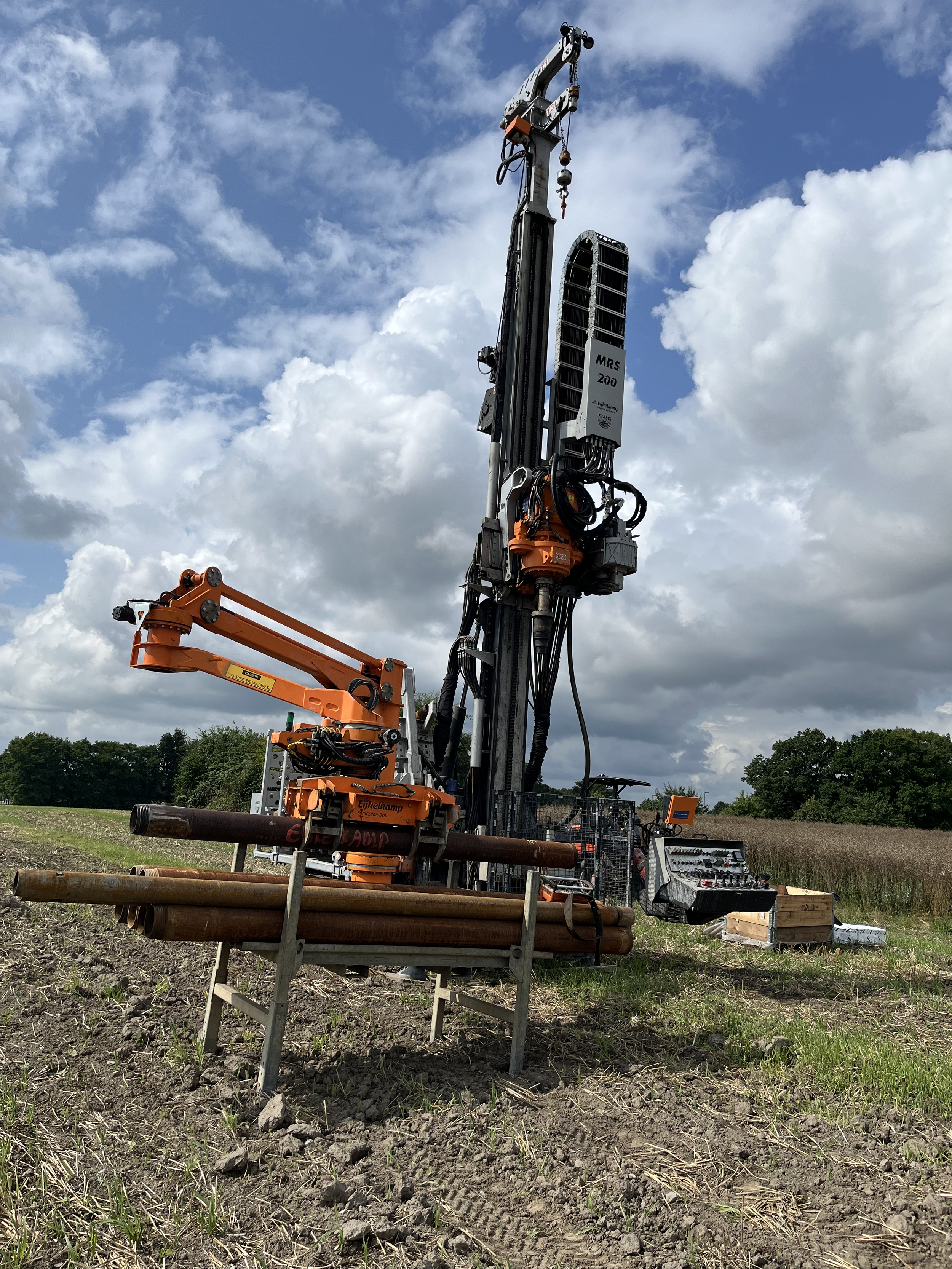 Ein Bohrgerät steht auf einer offenen, landwirtschaftlichen Fläche mit grünem Grasteppich im Hintergrund. Das Gerät ist schwarz und orange, mit verschiedenen hydraulischen Armen und Kontrollpanelen.