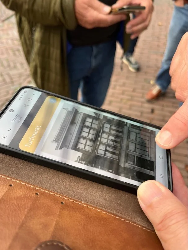 Persoon houdt smartphone vast die een zoekresultaat voor 'Tuinmarkt' toont met een afbeelding van een gebouw met ramen. Onderdeel van het Erfgoedspel.