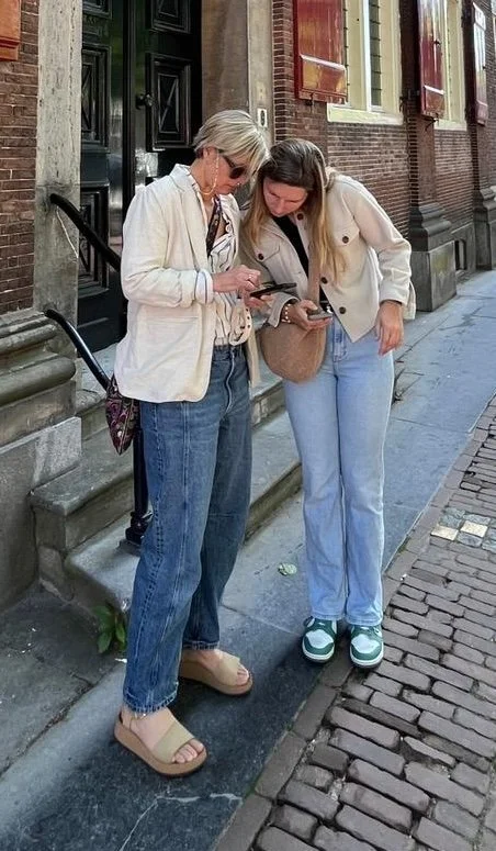 Twee vrouwen staan op een stoep, kijken naar hun smartphones, en praten met elkaar over het oplossen van een interactieve puzzel.