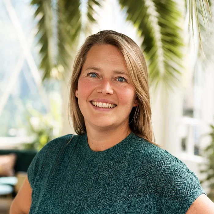Een lachende vrouw met schouderlange blond haar, gekleed in een groene trui, staand in een lichte, groene omgeving met palmbladeren op de achtergrond.