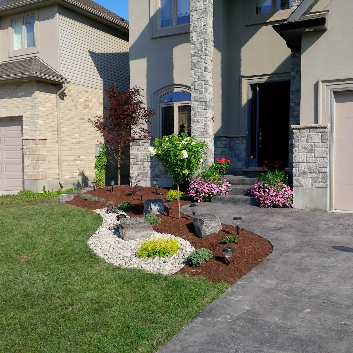 Front yard with a landscaped garden featuring a small tree, colorful flowers, decorative rocks, and pathway lighting in front of a modern house with stone accents and a porch.