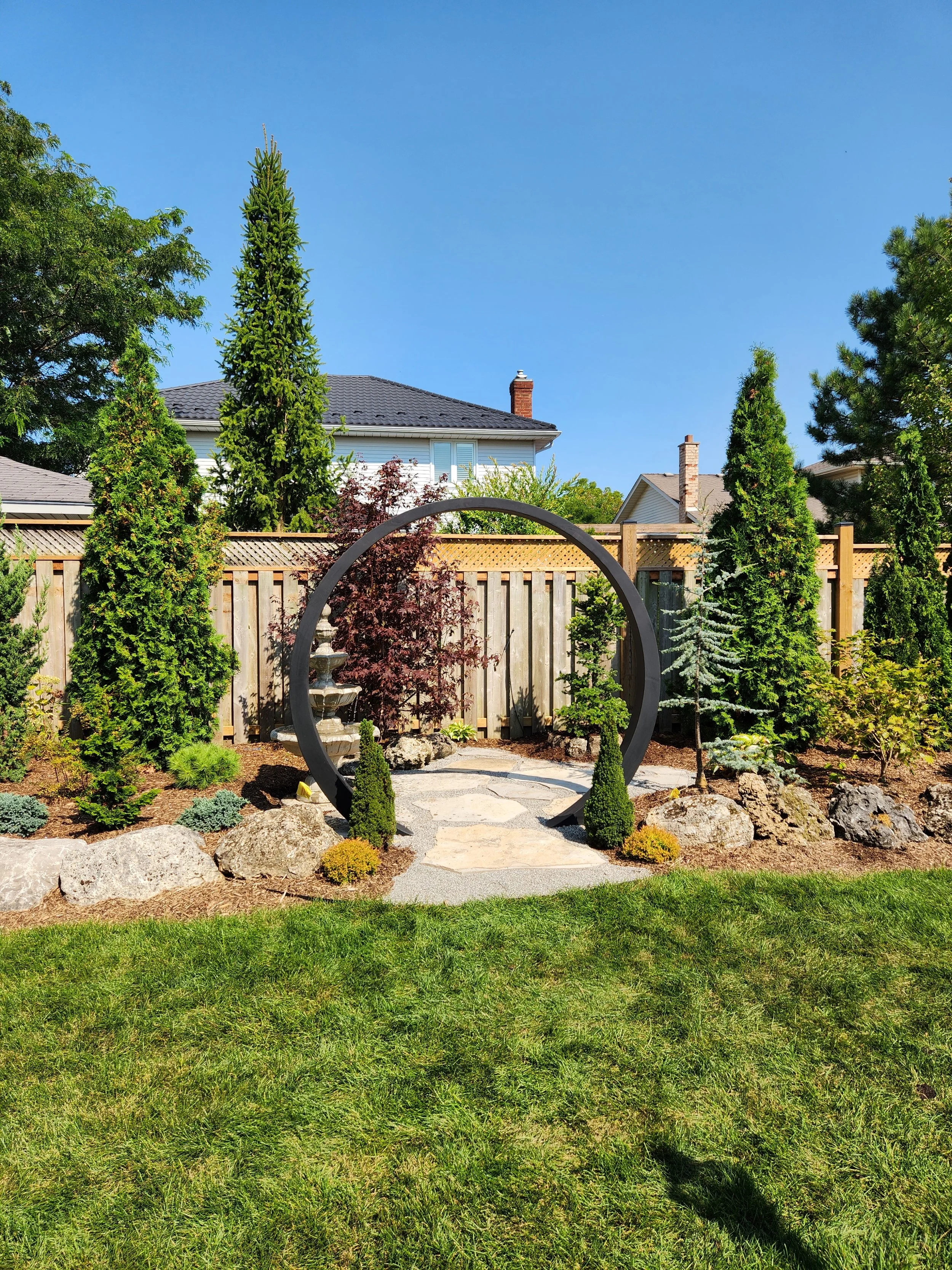 A landscaped backyard garden with a curved metal archway, moon gate, a stone pathway, various small trees, bushes, large rocks, and a wooden fence