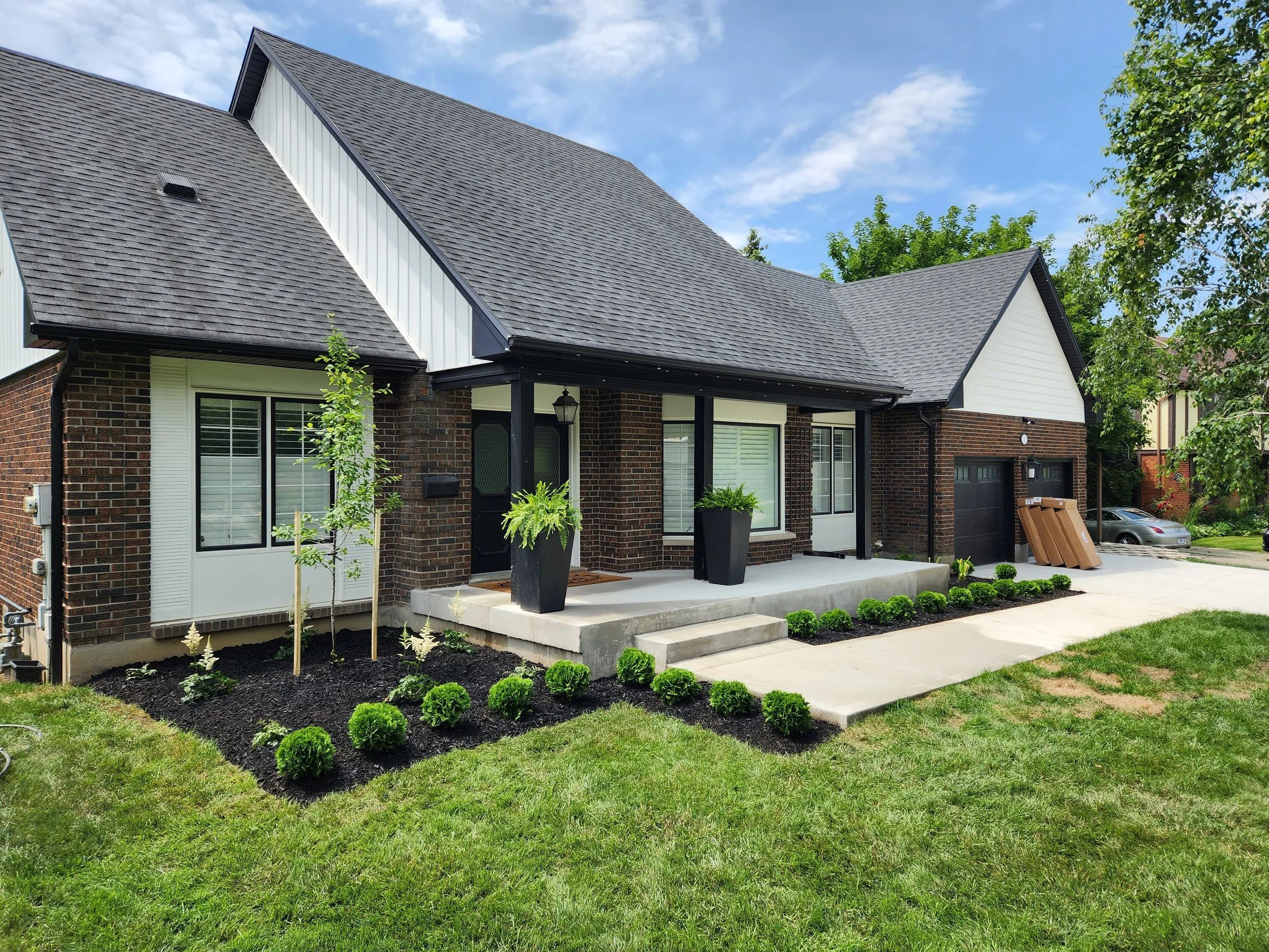 Front yard of a brick house with a concrete porch and black planters, landscaped with small bushes and young trees, on a sunny day with a blue sky.