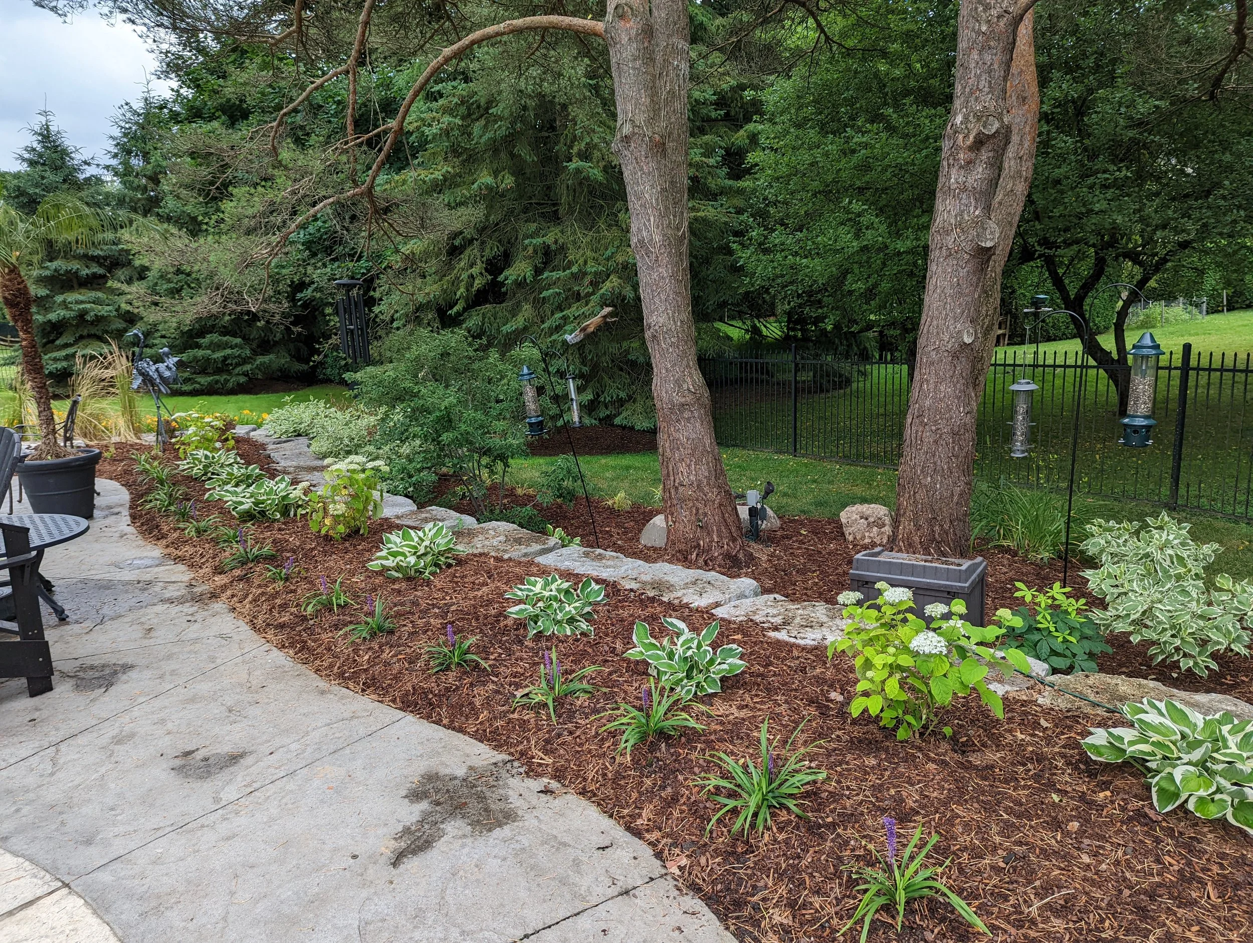 A landscaped backyard garden with a curved stone path, a tree with hanging birdfeeders, flowering plants, decorative garden sculptures, and a black metal fence in the background.