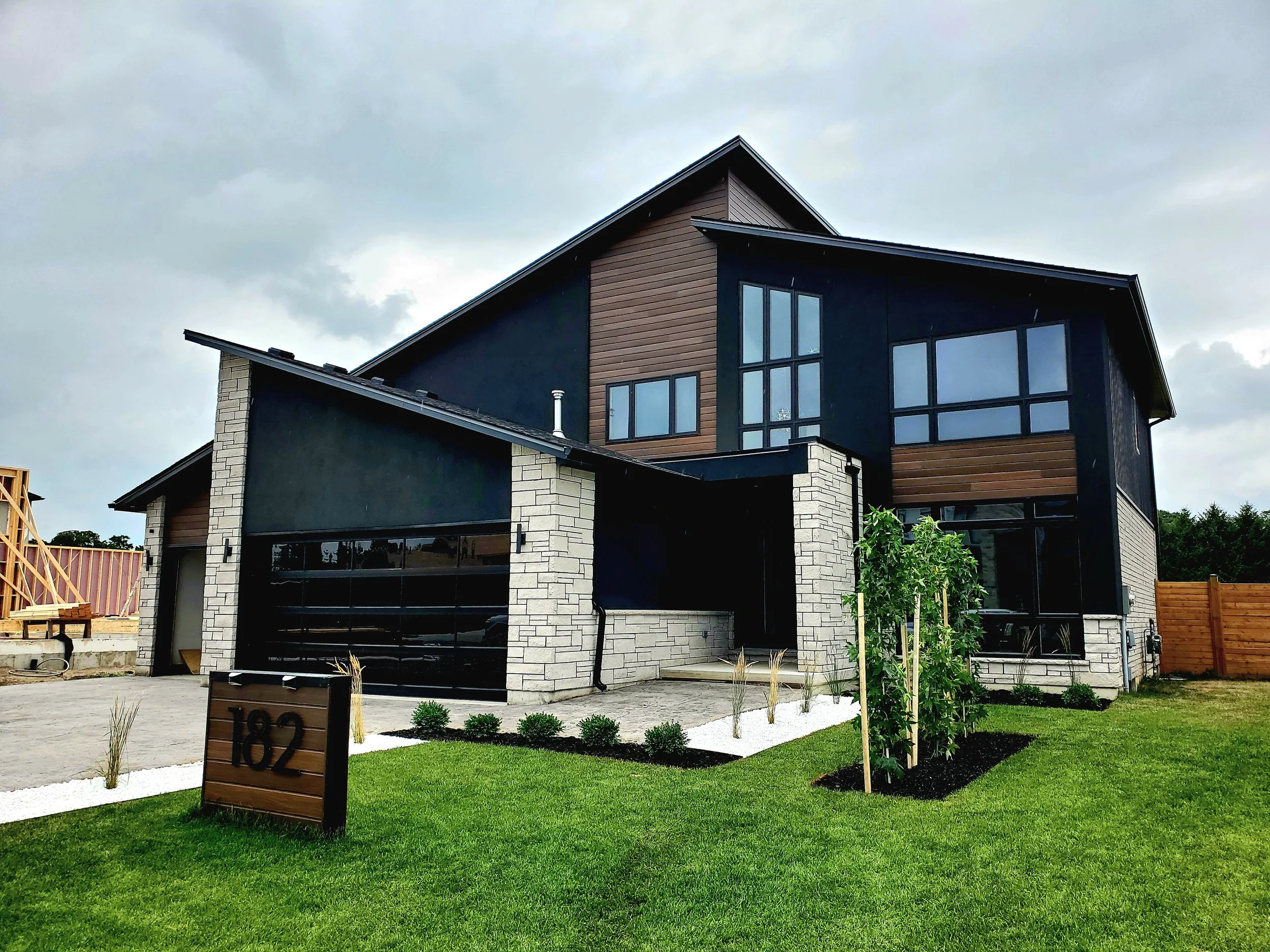 Modern two-story house with black and wood siding, large windows, a landscaped front yard with green grass, small shrubs, and a wooden house number sign showing 182