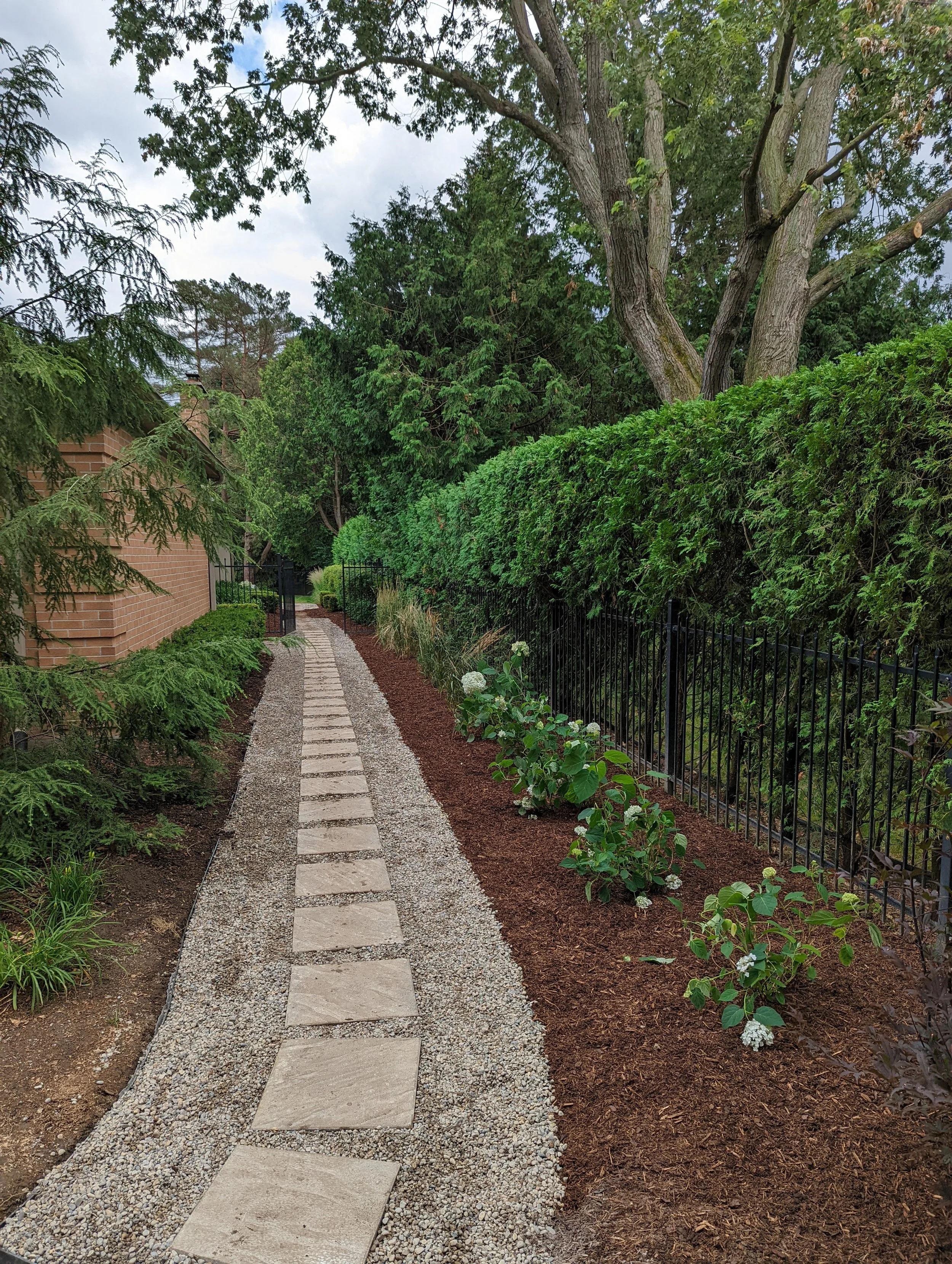 A winding stone pathway bordered by green plants and shrubbery, with a brick building on the left and a black metal fence on the right, set among tall trees with cloudy sky overhead.