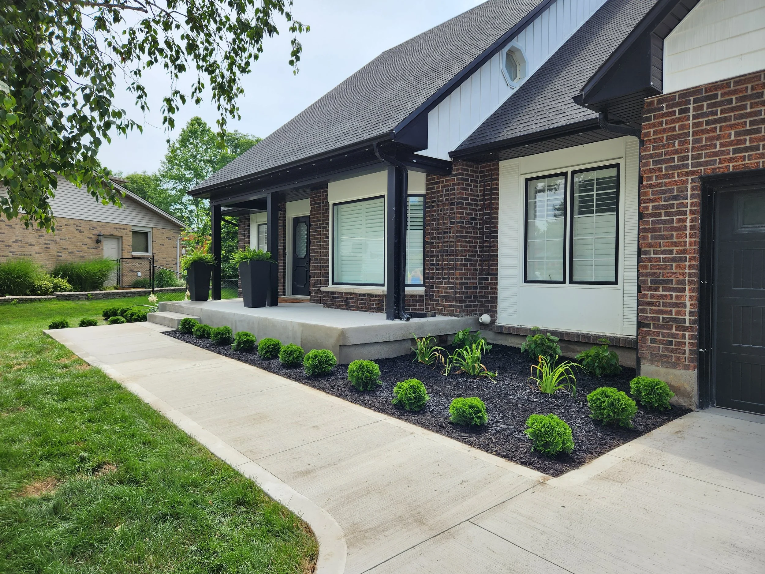 Front porch of a house with black door, white window, small garden, and concrete steps.