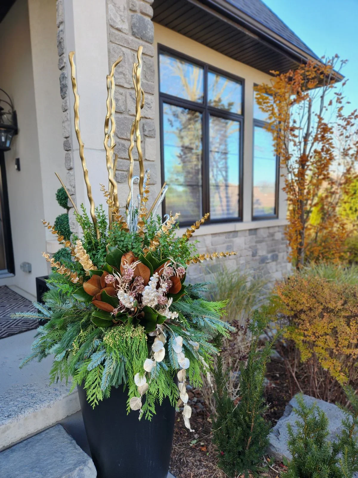 A decorative floral arrangement with green foliage, white and pink flowers, and tall gold and silver twisted branches in a black vase outside a house with large windows and stone siding. Winter planter, cedar, pine, spruce, magnolia, berries, corkscr