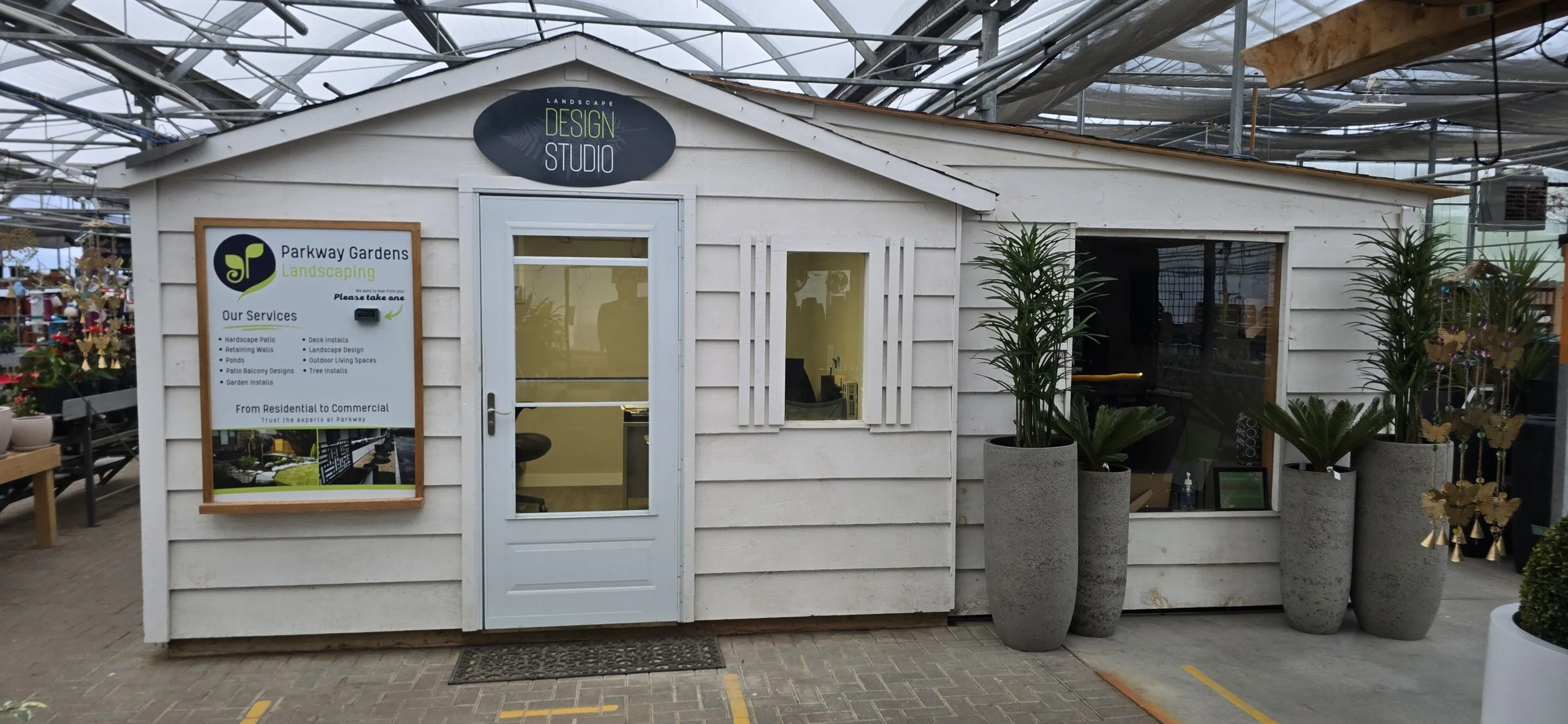 A small white wooden building labeled 'Landscape Design Studio' with large planters of tall green plants in front. To the left, a sign displays services of Parkway Gardens Landscaping, including lawn and patio designs. The building is in an outdoor setting, possibly a garden center or nursery, with glass roof panels overhead.