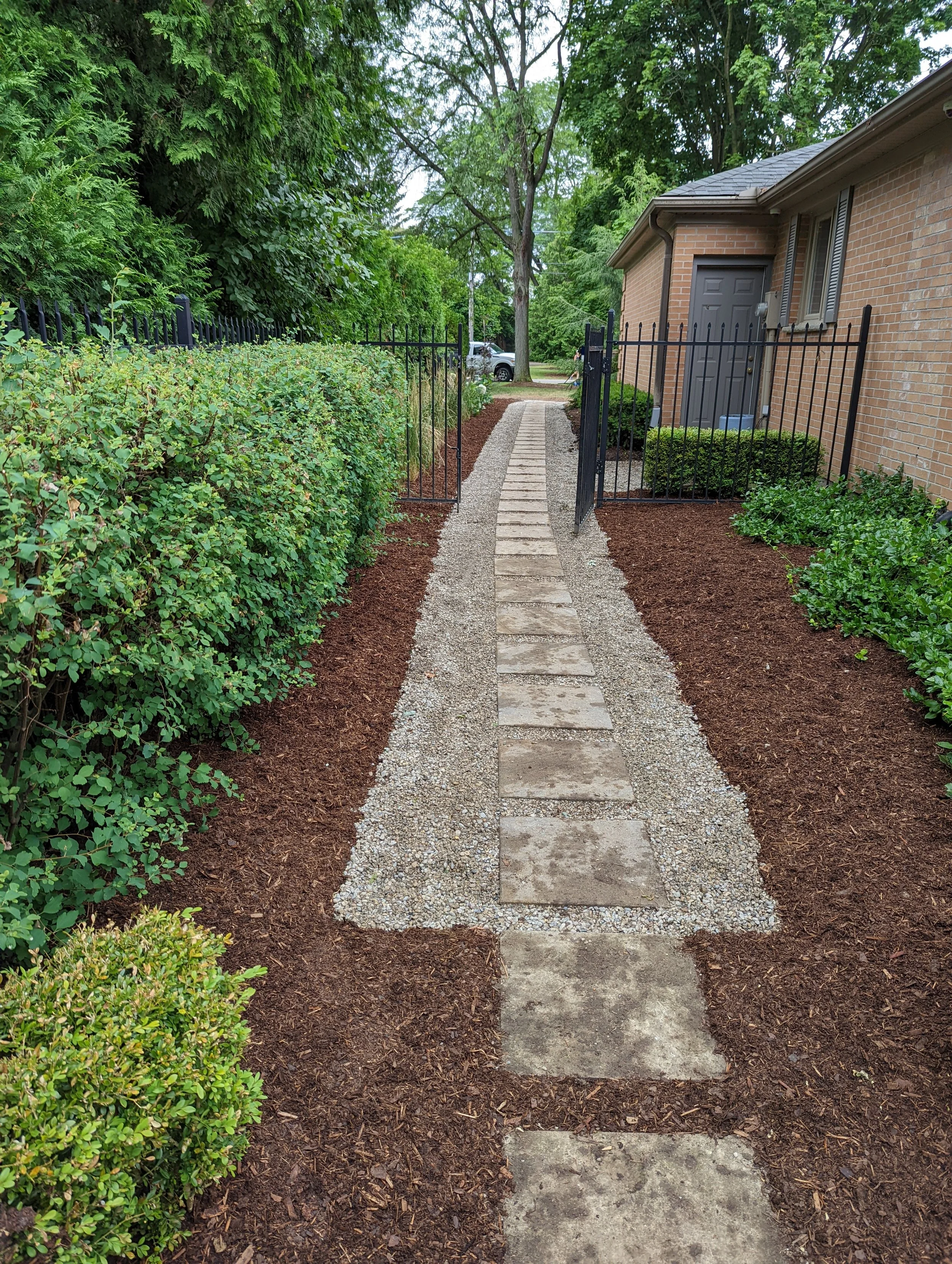 A narrow stone pathway with rectangular pavers runs through a landscaped garden, bordered by mulch and shrubbery, leading up to a gated entrance near a house with brick walls and a closed door. Tall trees provide greenery overhead.