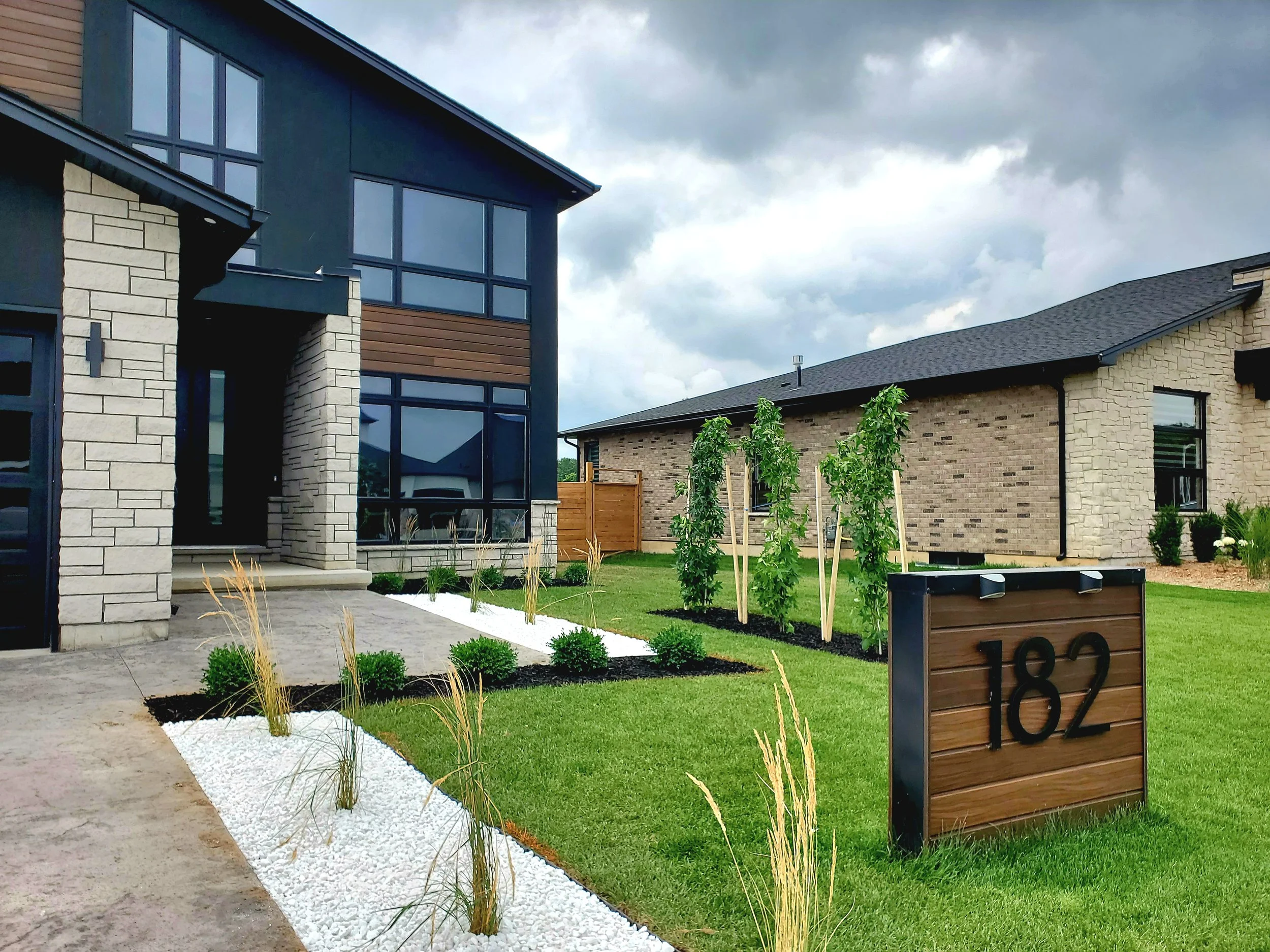 Modern residential house with large windows, well-maintained lawn, and landscaped garden featuring small bushes and tall grasses, numbered 182 on a wooden sign.