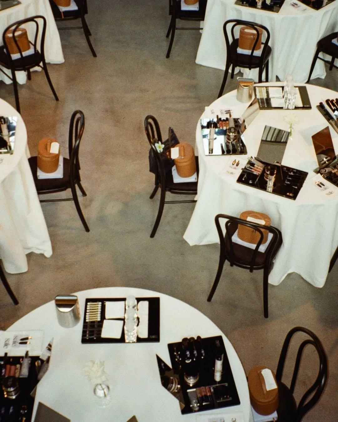 Round tables with white tablecloths set up for a makeup or beauty event, each with makeup products and tools, chairs arranged around. Small decorative flower arrangements on the tables.