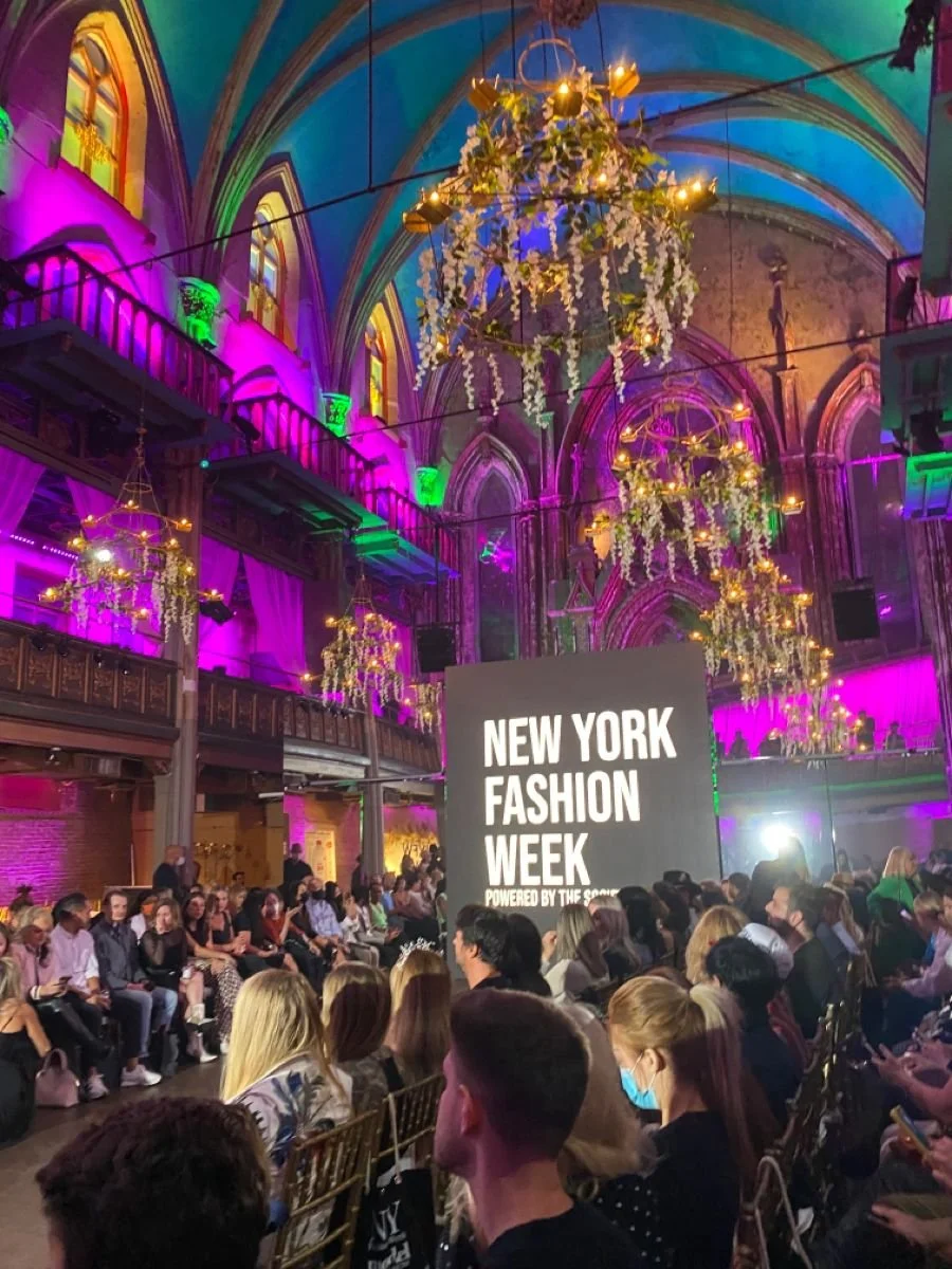 Audience seated at New York Fashion Week event inside a large, ornate hall with colorful lighting, chandeliers, and flower decorations hanging from the ceiling.