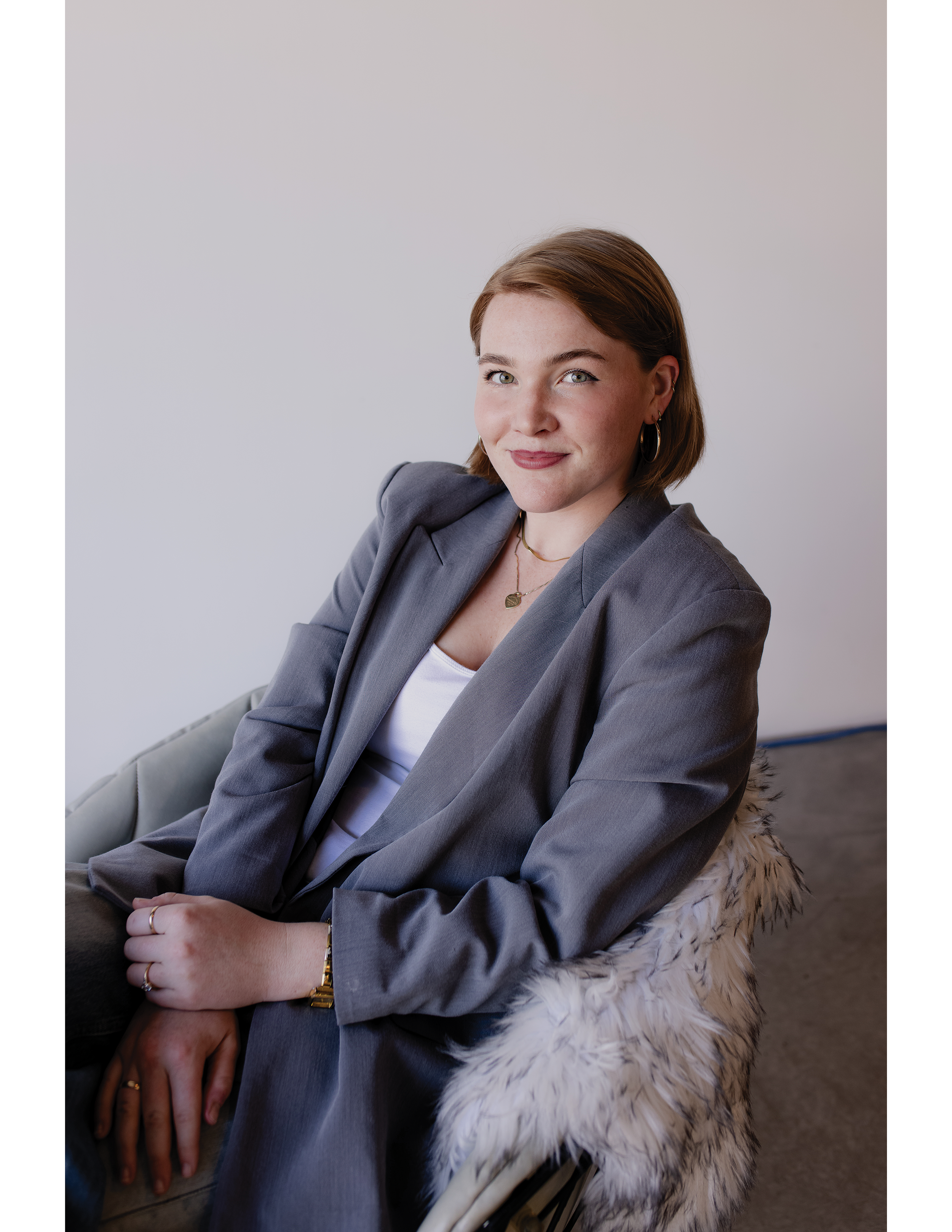 A young woman with brown hair, wearing a gray blazer over a white top, sitting on a beige chair with a fluffy white pillow, smiling softly at the camera.