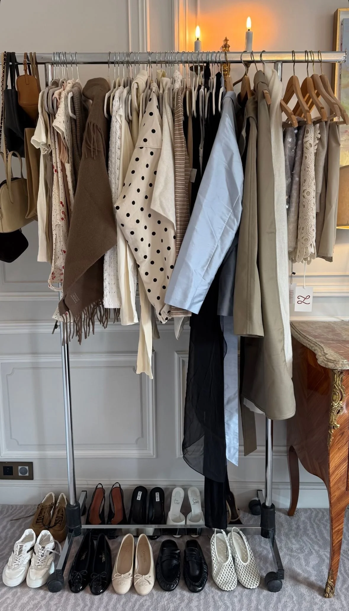 A clothing rack with various beige, white, black, and patterned garments hanging on wooden and plastic hangers, with shoes arranged underneath and a vintage fireplace with candles in the background.