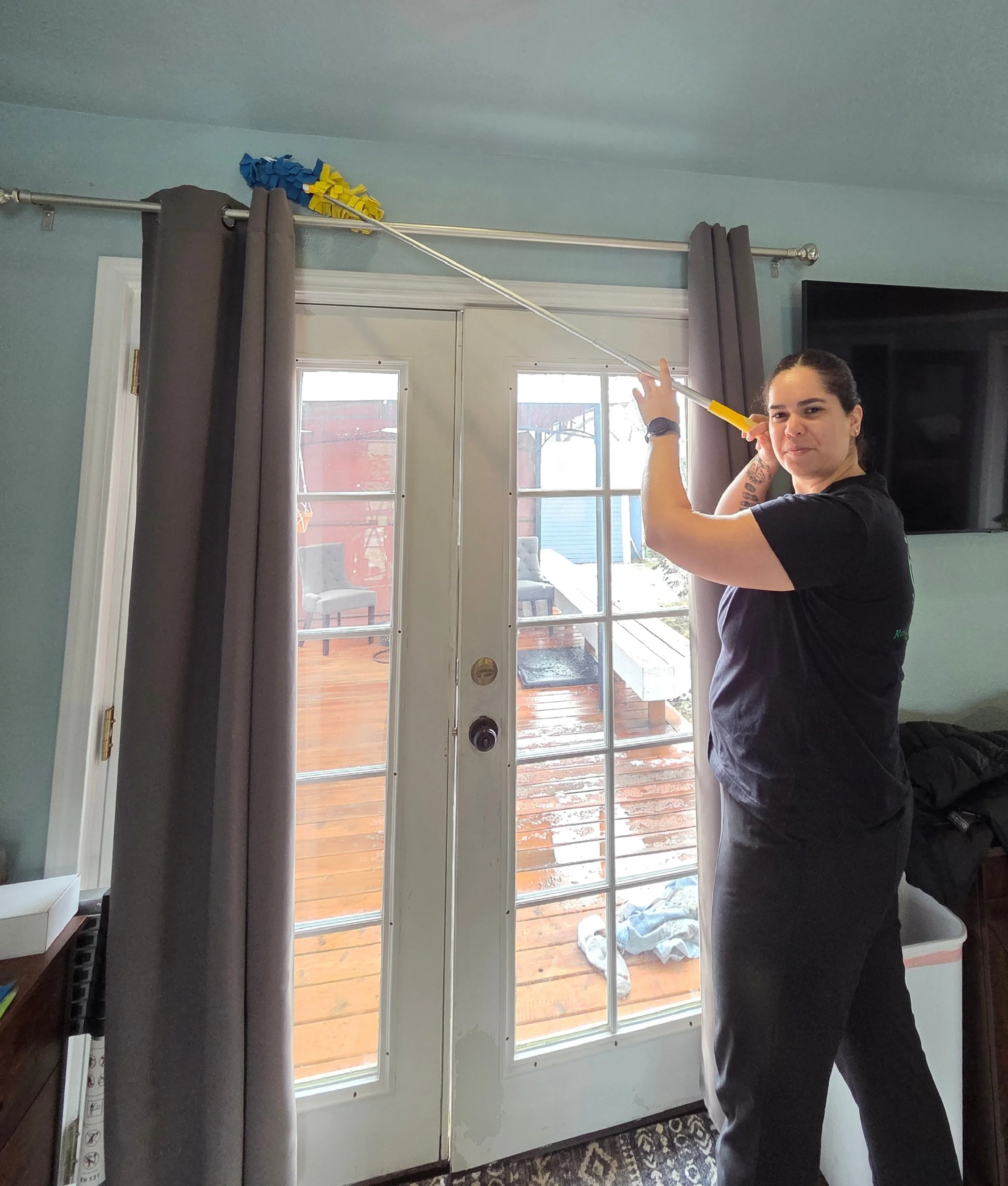 A woman is using a long-handled duster to reach the top of a curtain rod above a glass door, with an outside deck visible through the door.