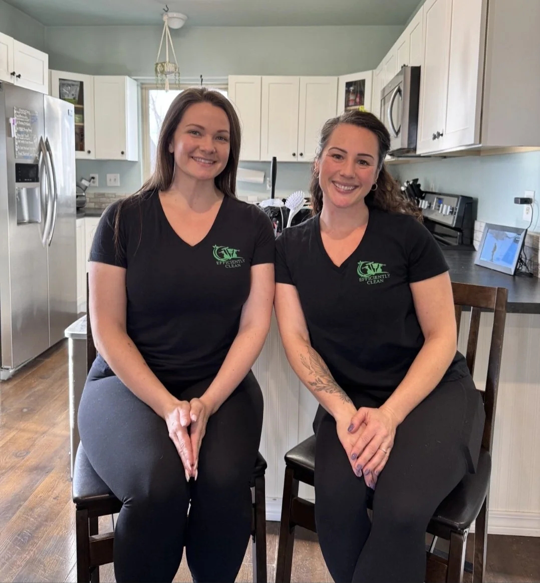 Two women sitting on chairs in a kitchen, smiling at the camera, wearing black shirts with a green logo that reads 'Efficiently Clean'.