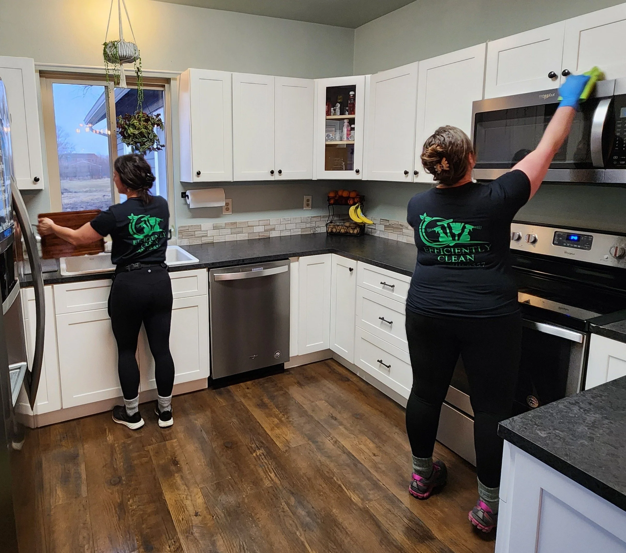 Two women cleaning a kitchen, one wiping the microwave and the other taking items from a cabinet, both wearing black T-shirts with green text and logos.