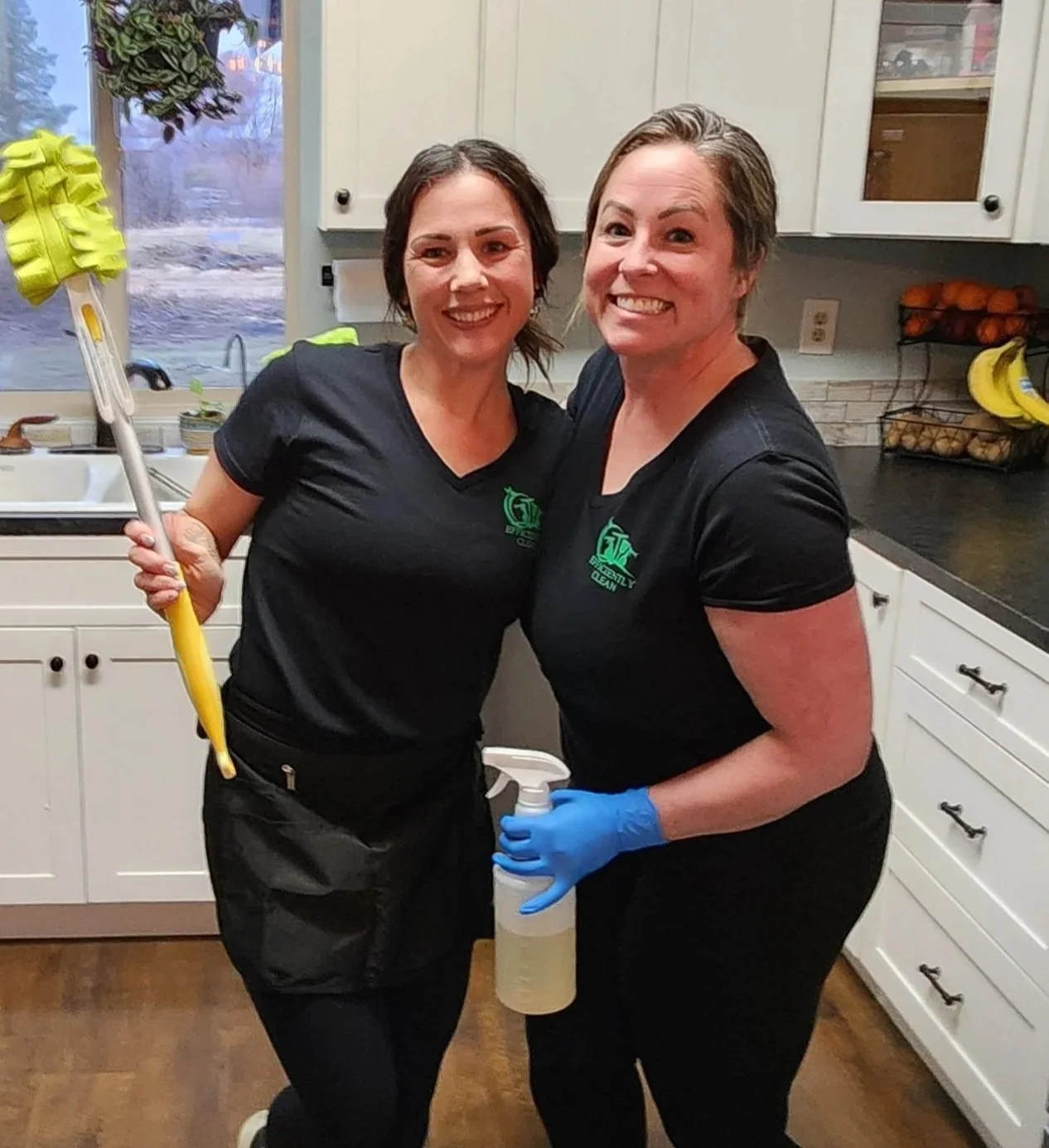 Two friendly women in smiling in the kitchen of one of their cleaning clients. One lady is holding a duster and the other is holding a cleaning product spray bottle.