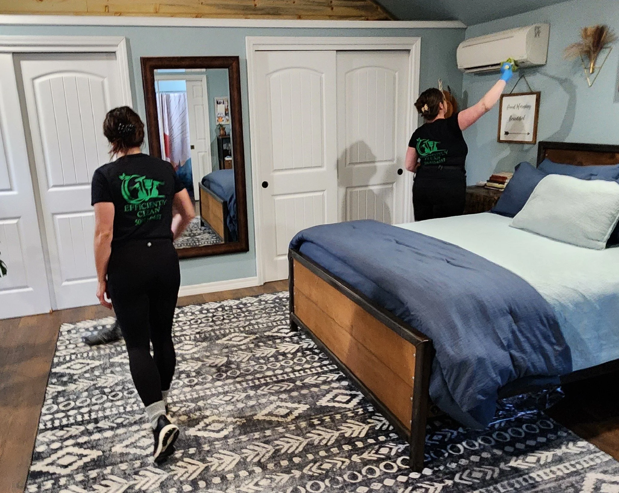Two women cleaning a bedroom, one is vacuuming the floor while the other is using a spray bottle to clean an air conditioning vent, with a bed, mirror, and framed sign on the wall in the room.