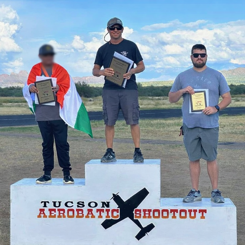 Three men standing on a winners' podium during the Tucson Aerobatic Shootout event, holding awards. The man in the center is on the top step, wearing sunglasses, a black T-shirt, cargo shorts, and a cap. The man on the left, with his face blurred, is wrapped in an Italian flag. The man on the right is wearing sunglasses, a gray T-shirt, and shorts. The background features a field and distant mountains under a partly cloudy sky.