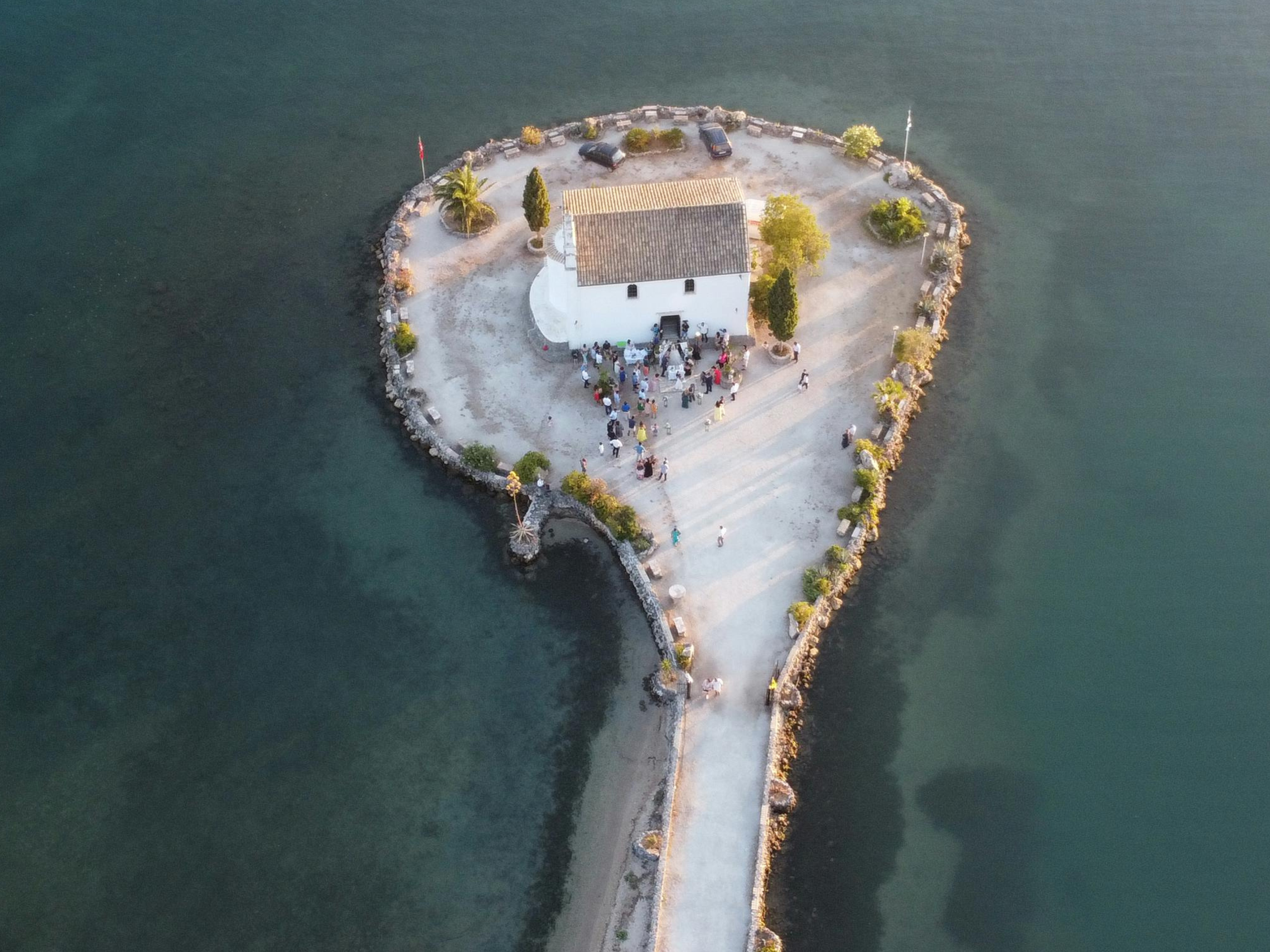 An aerial view of a small white church on a tiny island surrounded by water, with a pathway leading to the island and people gathered around the church entrance.