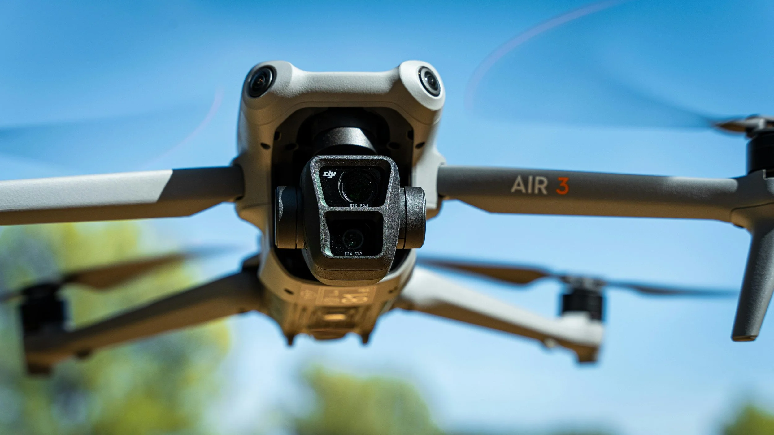 Close-up of a DJI Air 3 drone flying outdoors with blue sky and green trees in background.
