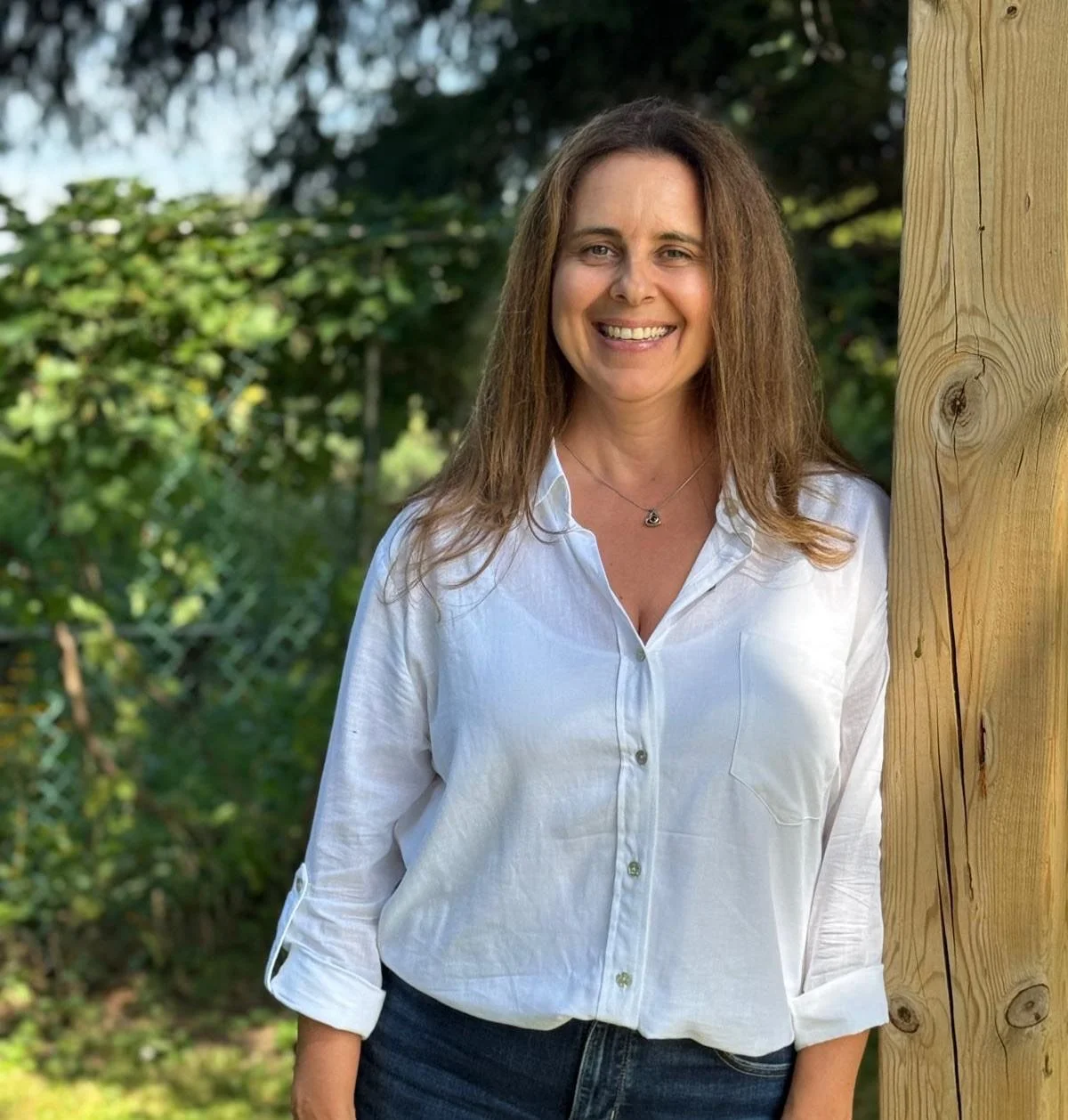 A smiling physiotherapist in Burlington Ontario with shoulder-length brown hair, wearing a white button-up shirt and blue jeans, standing outdoors next to a wooden post with greenery in the background.