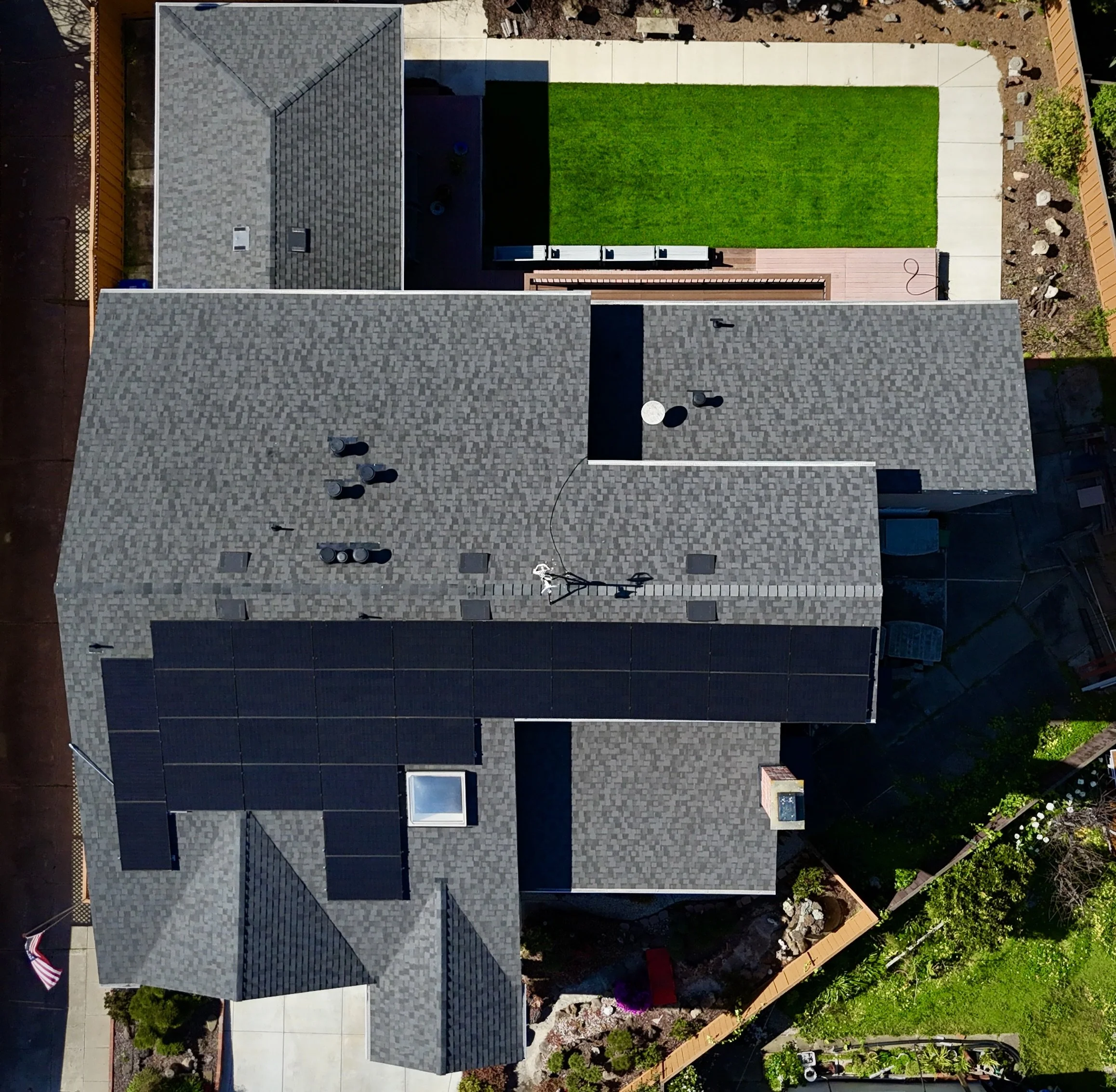 An aerial view of a house with solar panels on the roof, a backyard with a green lawn, a deck, and a paved area. There's a flagpole with an American flag and a garden on the side.