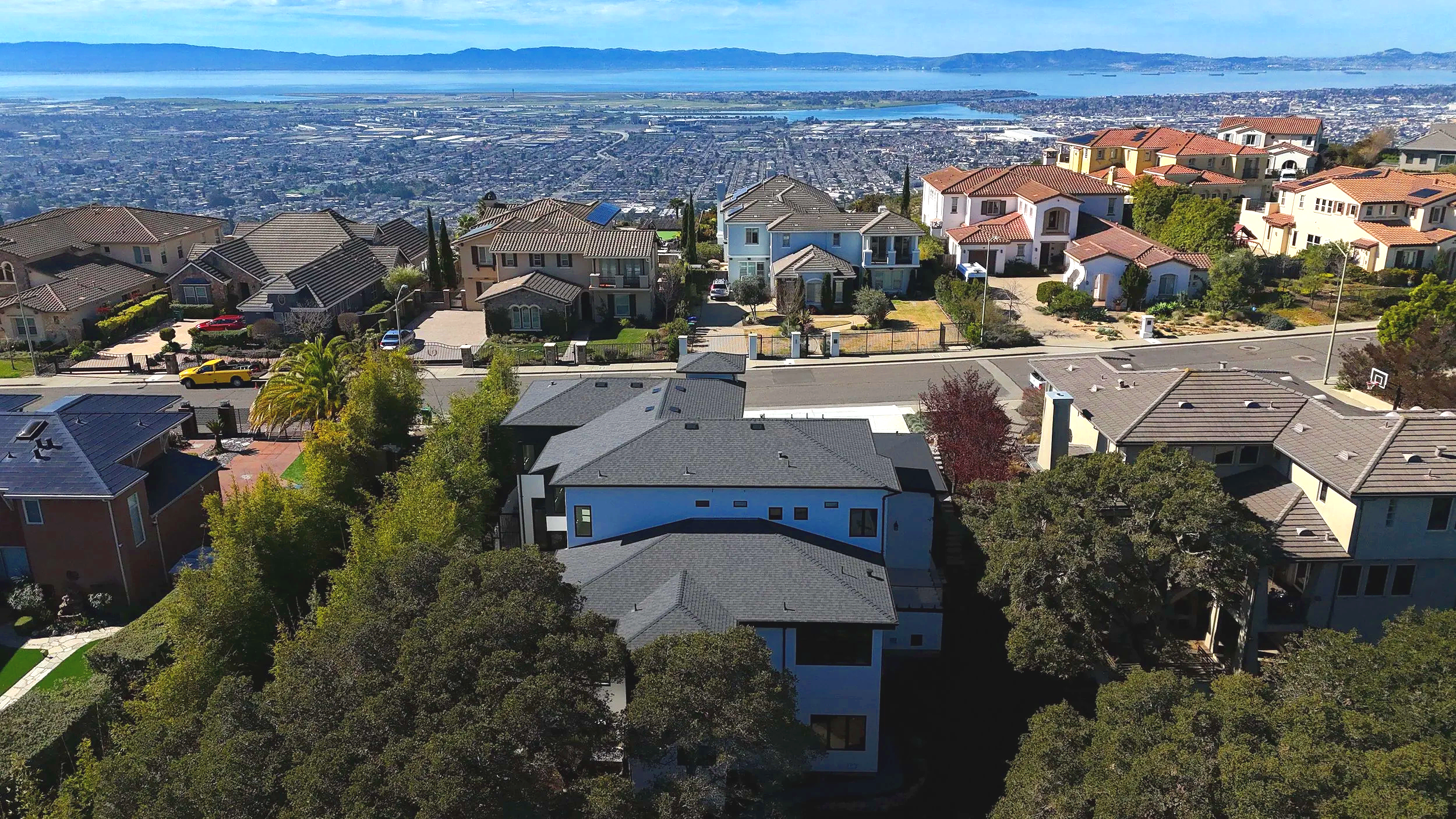 Aerial view of a neighborhood with modern houses, trees, streets, and a distant body of water under a clear blue sky.