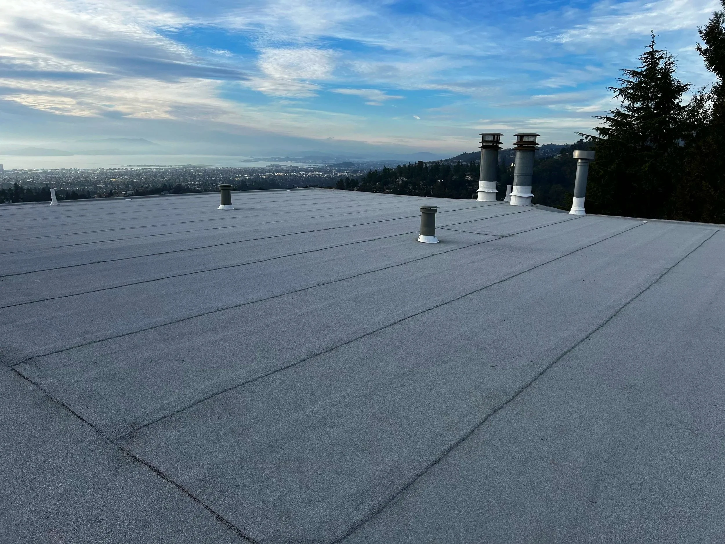 Flat rooftop with gray roofing material, multiple metal vents and chimneys, overlooking cityscape and ocean in the distance, with a partly cloudy sky.