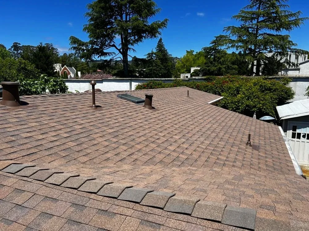 View of a rooftop with asphalt shingles, vent pipes, and a skylight, surrounded by trees and neighboring houses under a clear blue sky.