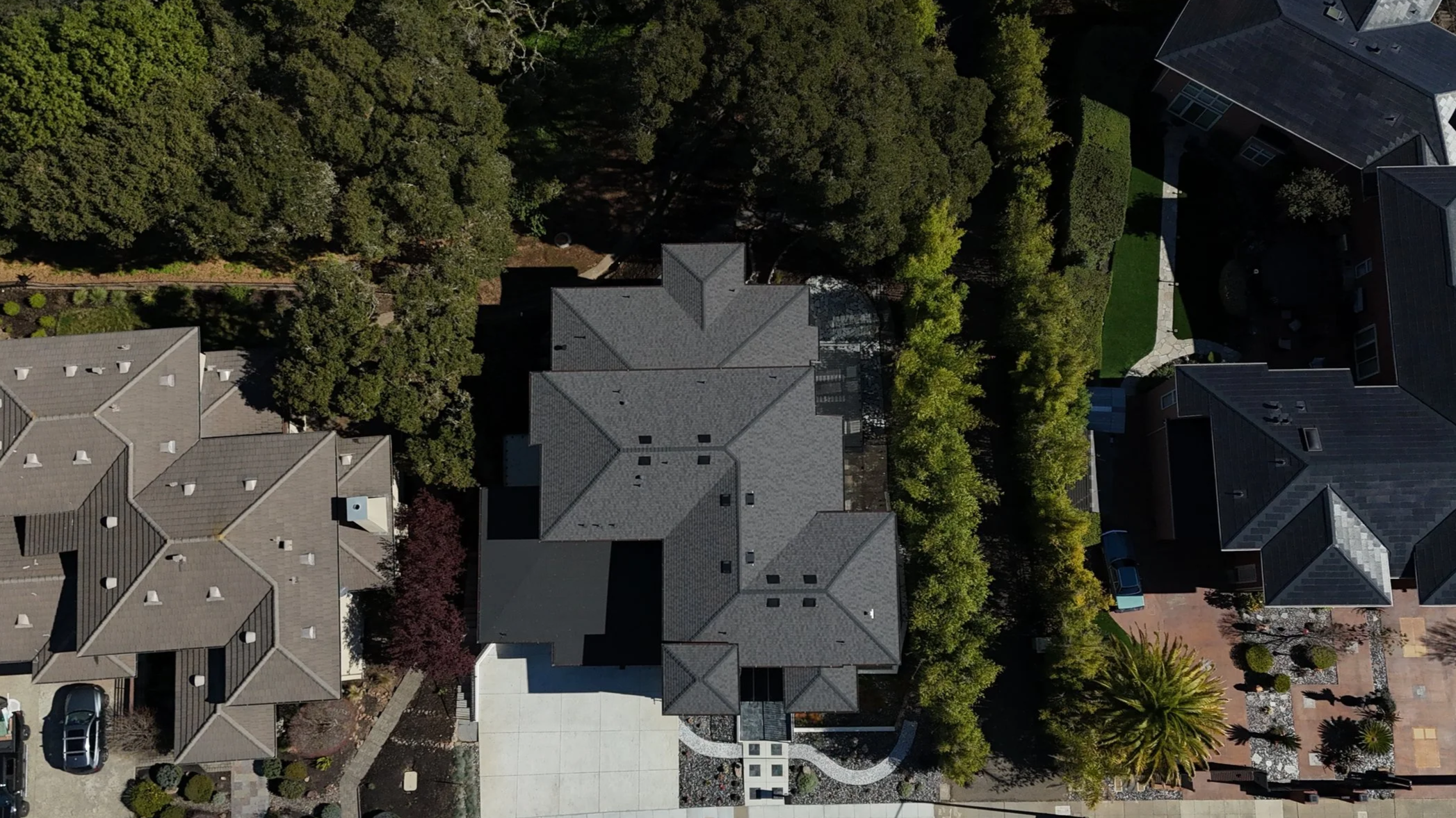 An aerial view of several houses with gray roofs, lush green trees, a well-maintained lawn, and a driveway with parked cars.