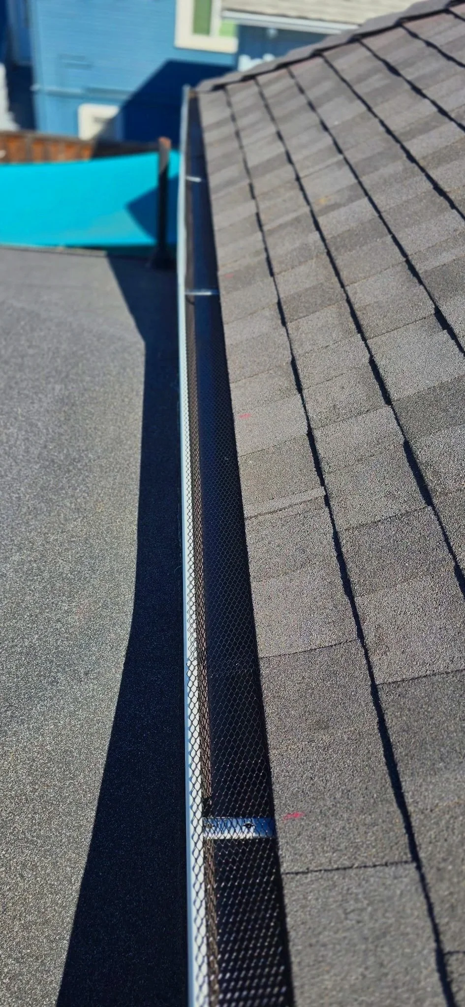 Close-up of a roof gutter with shingles on the roof, a section of the roof, and a neighboring blue house in the background.