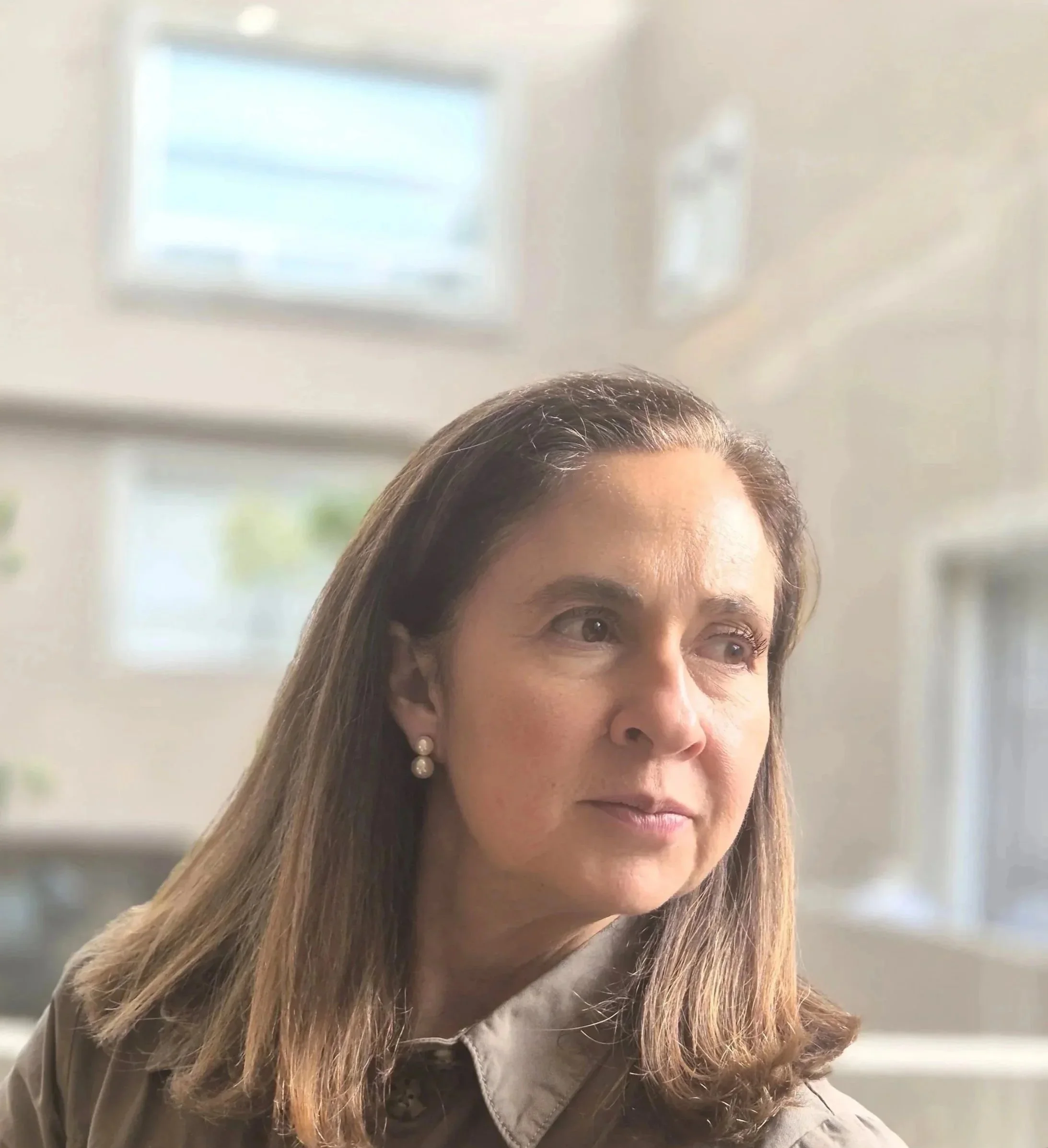 A woman with shoulder-length brown hair and pearl earrings looking thoughtfully to the side in a bright, modern indoor space with large windows.
